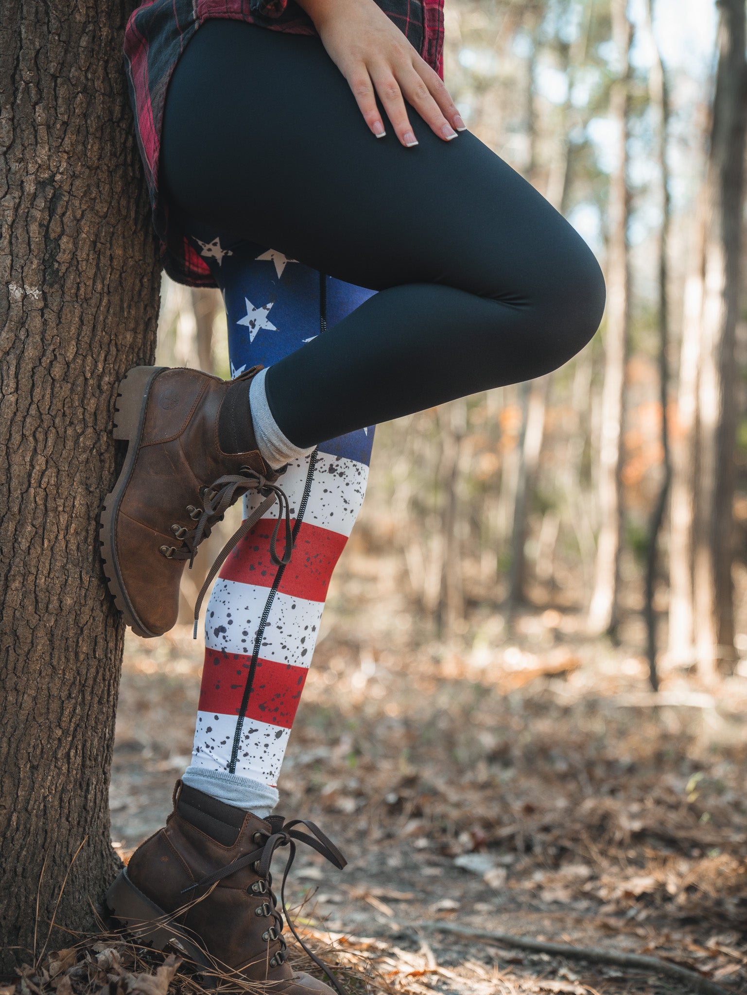 Amid fallen leaves and towering trees, a person stands by a tree, sporting American Savage Leggings from Born United Private Label beneath black pants. They wear a plaid shirt and brown boots, highlighting their patriotic style in the woods.
