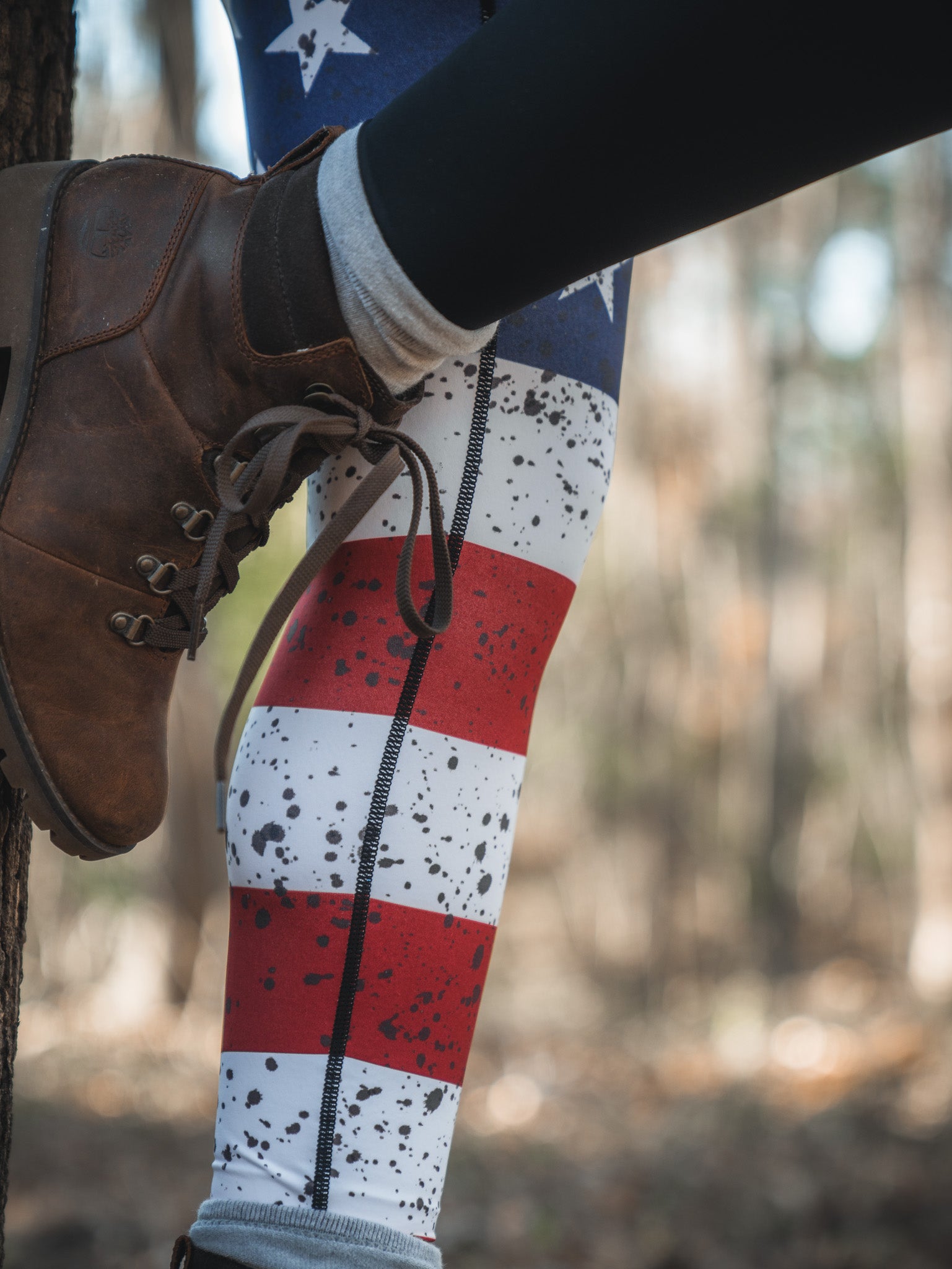 Wearing American Savage Leggings by Born United Private Label, featuring stars, stripes, and a splatter of red, white, and blue, a person in brown hiking boots rests their foot on a tree trunk within a lush forest.