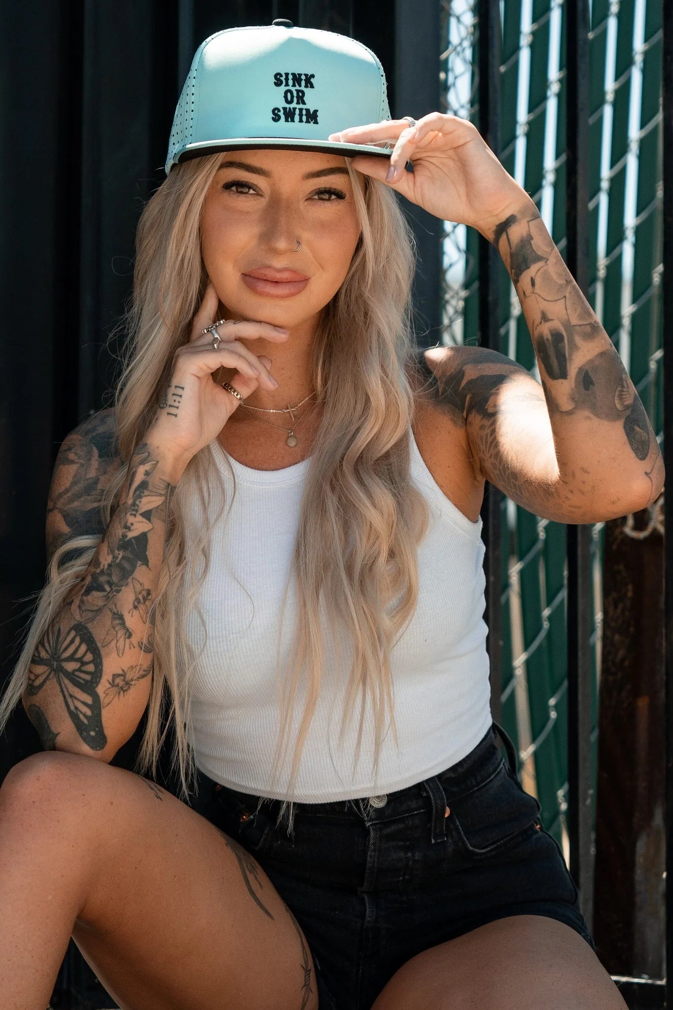 A tattooed individual with long hair, dressed in a white tank top and black shorts, sits outdoors smiling while holding the brim of a Sink or Swim Seafoam PVC Snapback by Relentless Betrayal. A fence and shadows feature in the background.