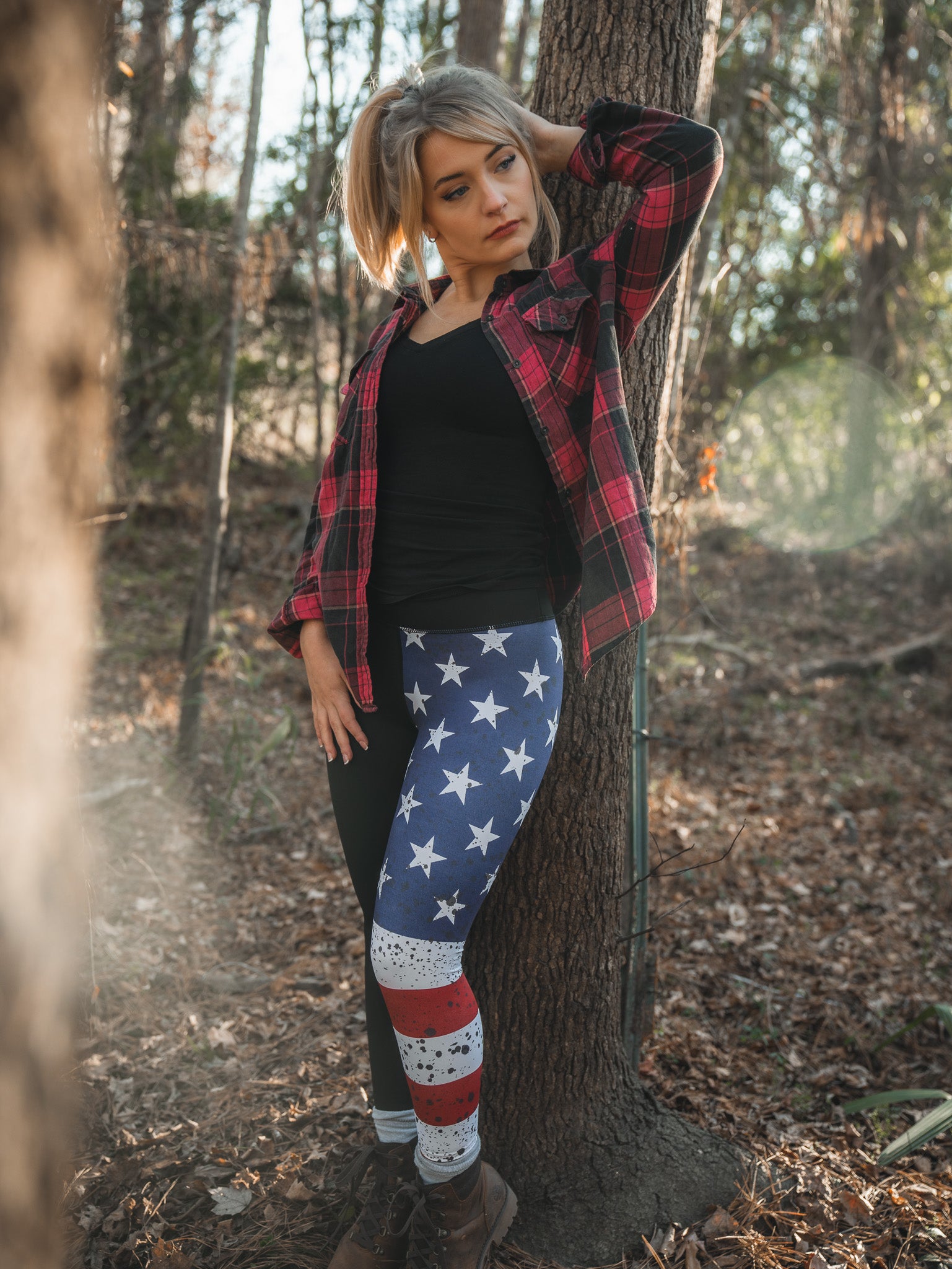 A woman wearing American Savage Leggings by Born United Private Label and a red plaid shirt leans against a sunlit forest tree, looking relaxed and contemplative with one hand behind her head and the other near her hip.