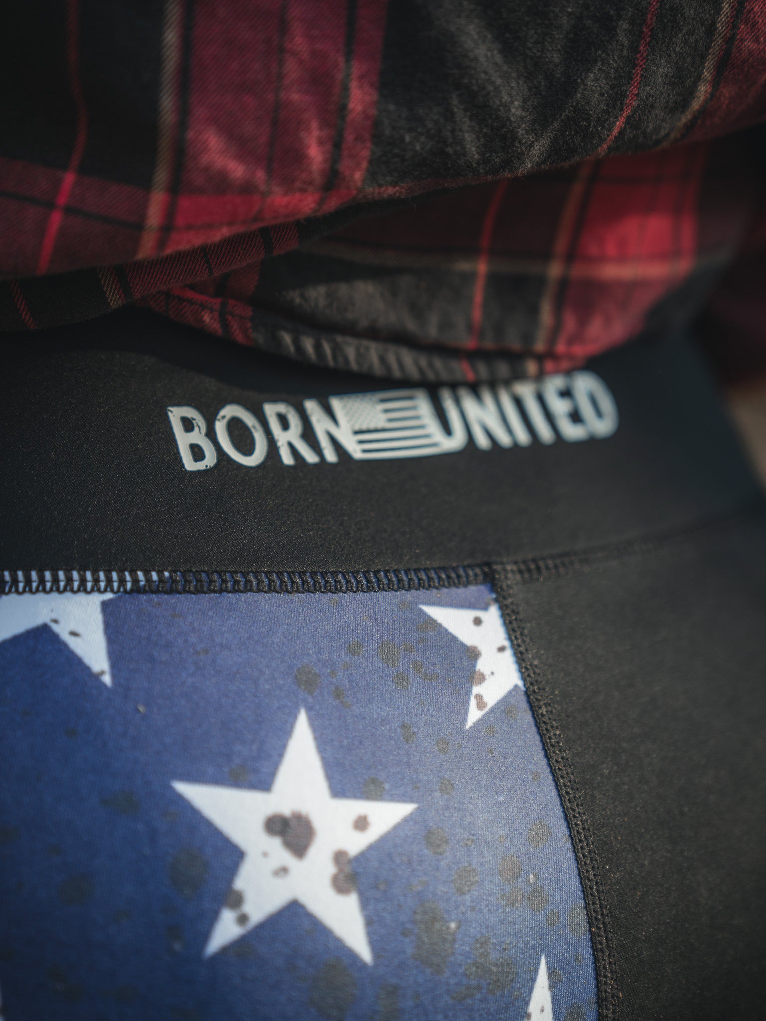 Close-up of someone wearing American Savage Leggings by Born United Private Label, featuring a stars and stripes pattern. BORN UNITED is emblazoned on the waistband with an American flag design in the letter R, paired with a red and black plaid shirt.