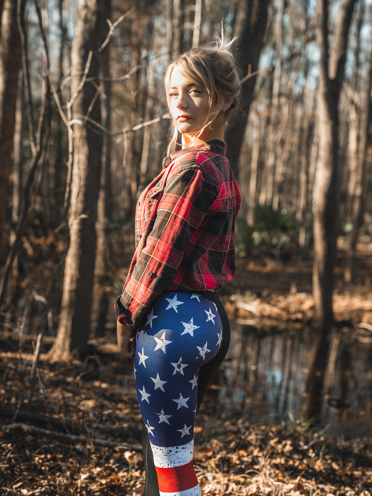 A person stands in a forest wearing Born United Private Labels red plaid shirt and patriotic American Savage Leggings, adorned with stars and stripes. Sunlight filters softly through the trees, casting shadows on the forest floor.