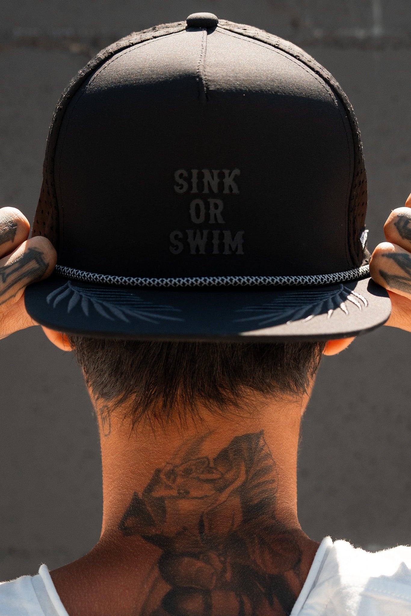 A tattooed individual adjusts their Sink or Swim Blackout PVC Snapback by Relentless Betrayal, a water-resistant cap embodying a pirate lifestyle vibe, against a plain wall backdrop.