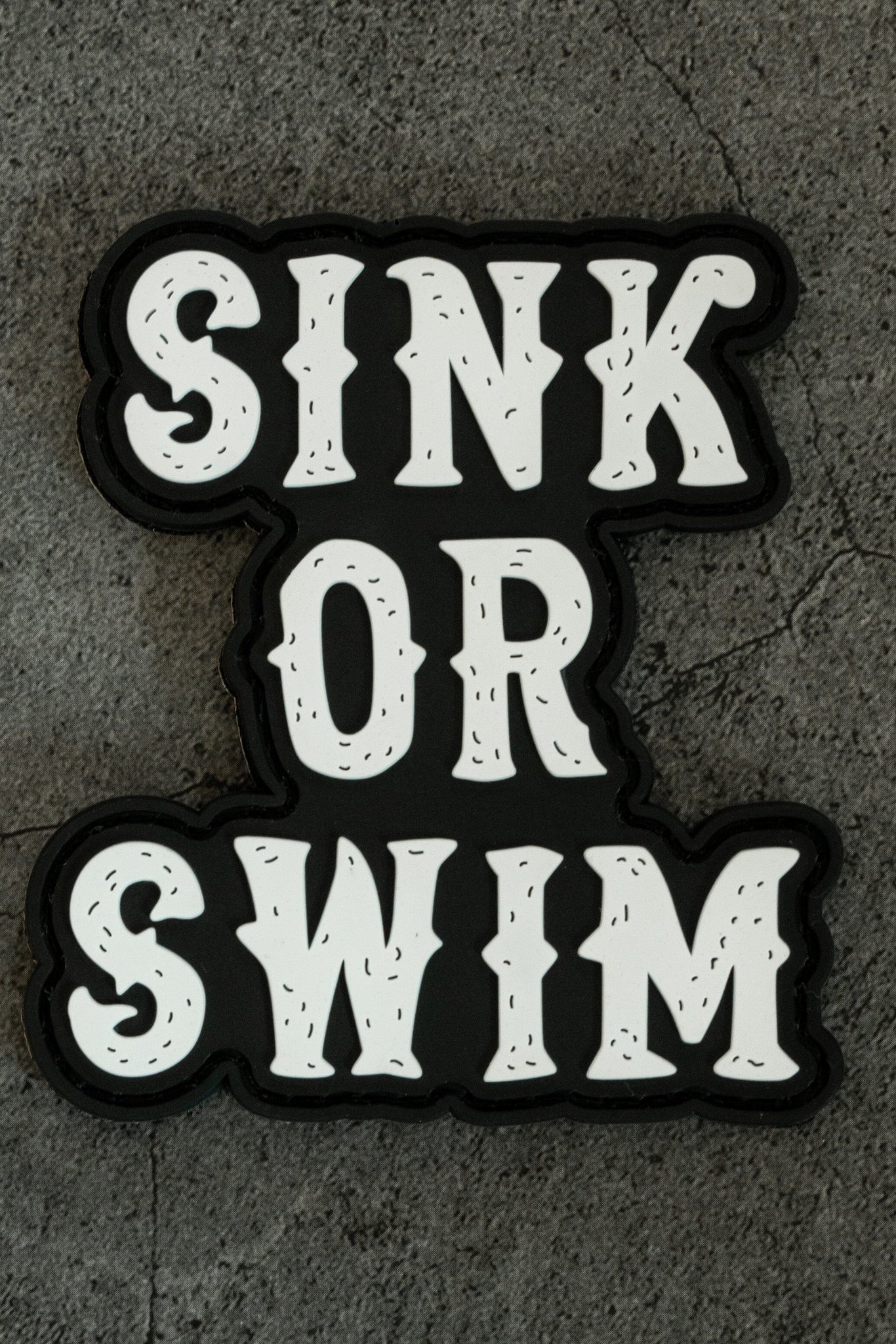 The Relentless Betrayals Sink or Swim Black and White PVC Patch features bold, white text on a dark ocean-like background, displayed on a textured gray surface.