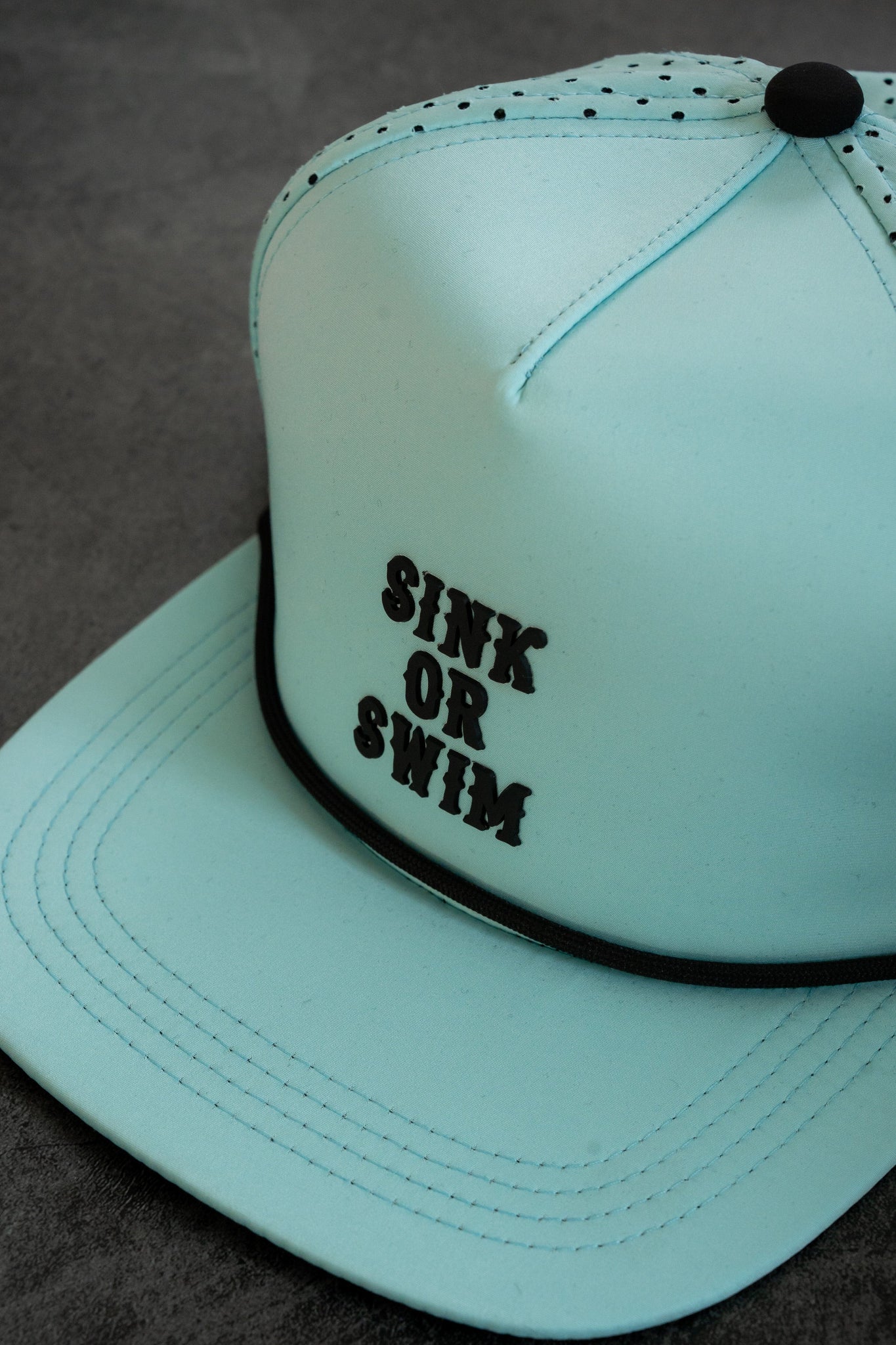 The Sink or Swim Seafoam PVC Snapback by Relentless Betrayal features black SINK OR SWIM text, a small black top button, black brim edging, and a sleek PVC rubber patch. It embraces the pirate lifestyle and sits stylishly on a gray surface.
