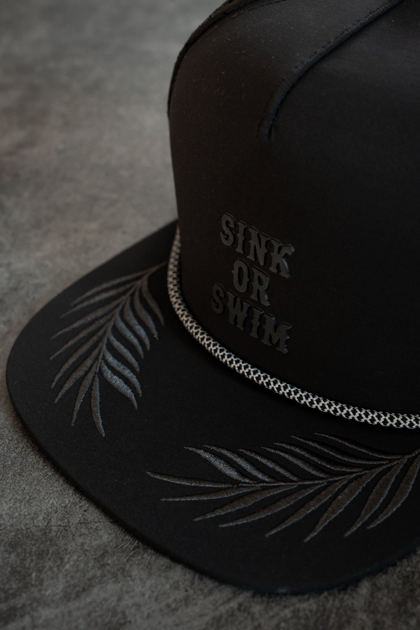The Sink or Swim Blackout PVC Snapback by Relentless Betrayal is a black cap styled for the pirate lifestyle with Sink or Swim on the front, palm fronds embroidered in gray on the brim, and a silver chain around the crown base, all resting on a textured gray surface to capture your adventurous spirit.