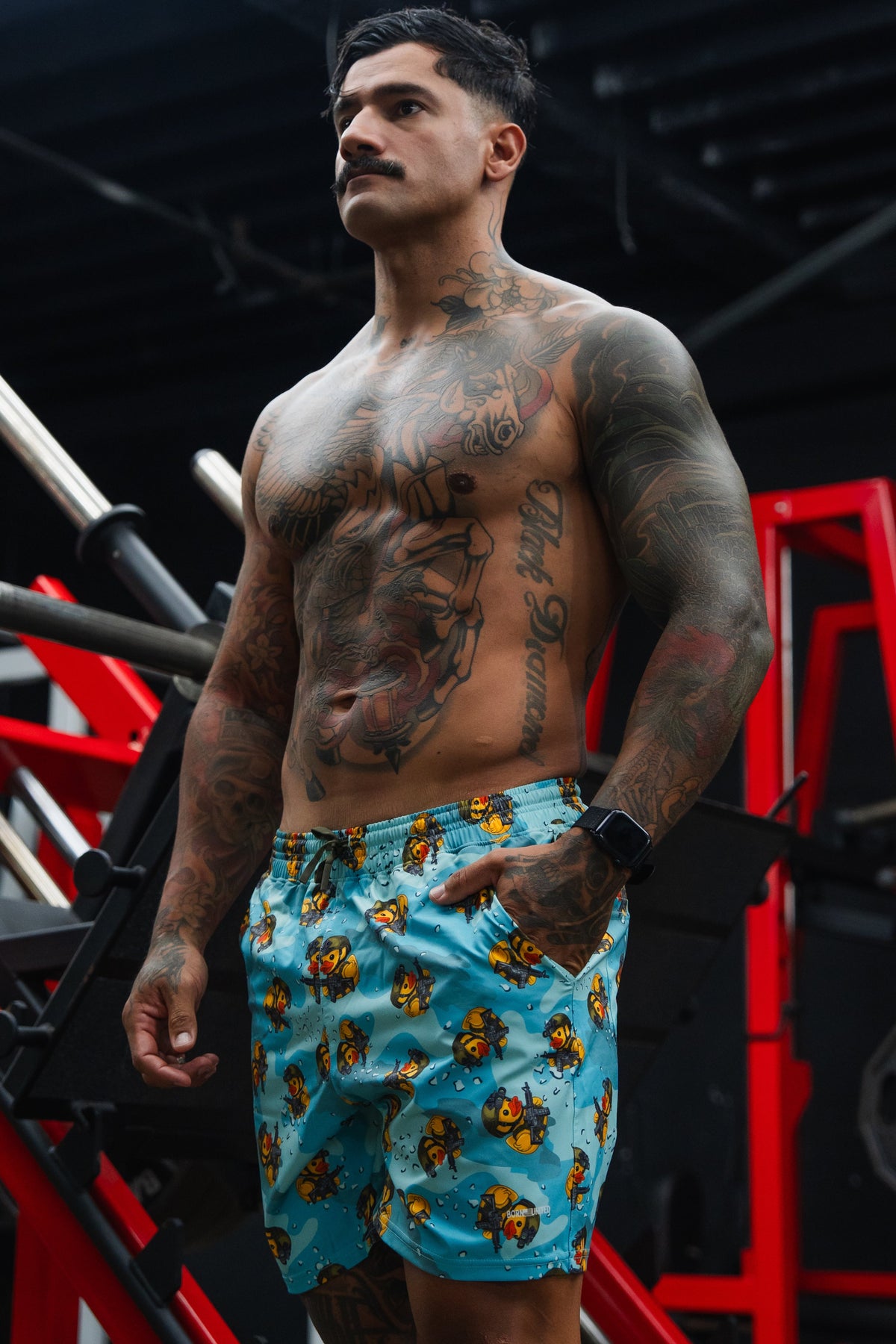 A muscular, tattooed man with a mustache stands shirtless in a gym, wearing Born United Private Label Men's Performance Hybrid Shorts 5.5" | TactiQuack. Red gym equipment and weight racks can be seen in the background.
