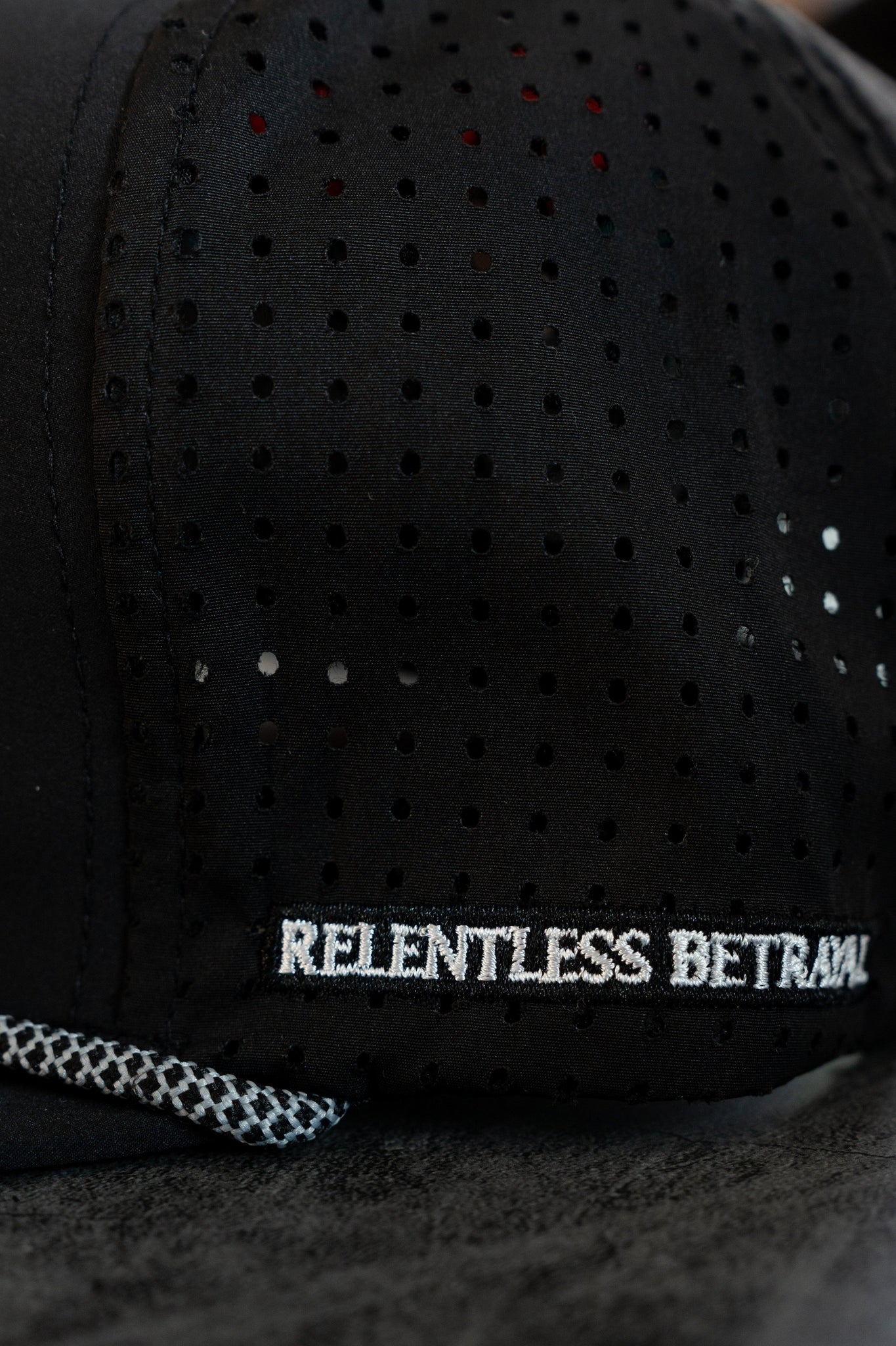 Close-up of the Sink or Swim Blackout PVC Snapback featuring a black cap with a perforated side design and textured grey edge. Relentless Betrayal is embroidered in white on the back, capturing the daring pirate lifestyle.