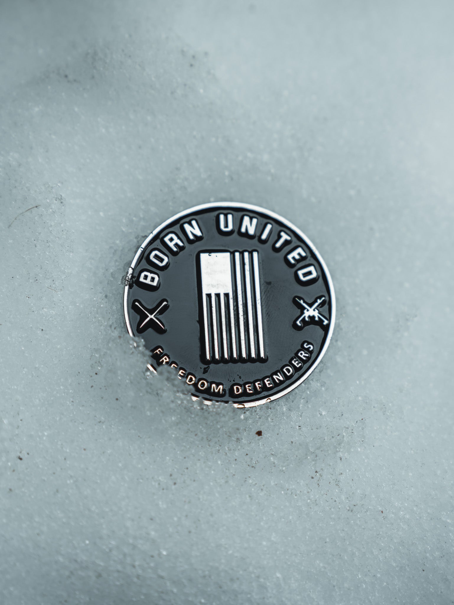A metallic Im Back Challenge Coin by Born United Private Label, partially embedded in snow, features Born United at the top and Freedom Defenders below. The design includes vertical stripes and two crossed tools.