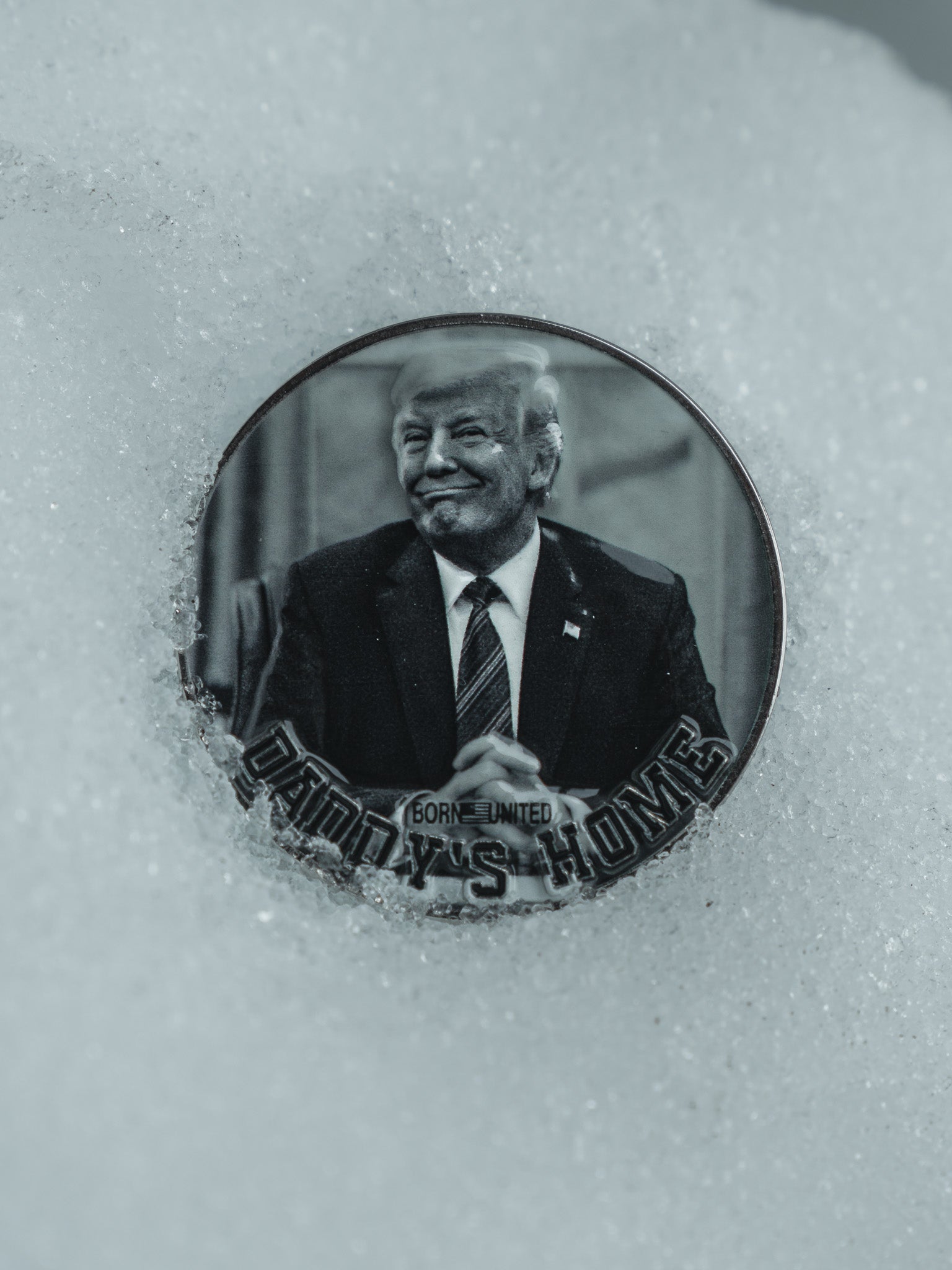 A round challenge coin from Born United Private Label rests partially in snow, displaying a man in a suit and tie with clasped hands. It reads DADDYS HOME and GONE UNITED, maintaining a monochromatic color scheme. Product: Daddy’s Home Challenge Coin.