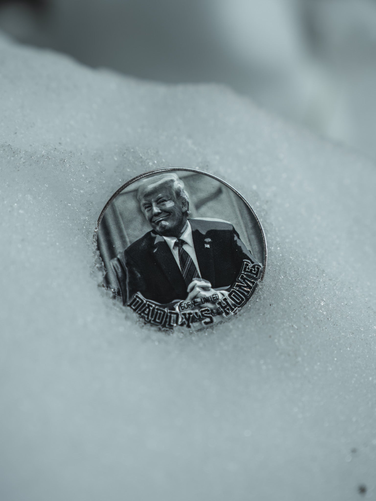 The Daddy’s Home Challenge Coin from Born United Private Label is a round pin with a grayscale image of a smirking person in a suit, partially embedded in snow, and the bold text DADDYS HOME at the bottom.