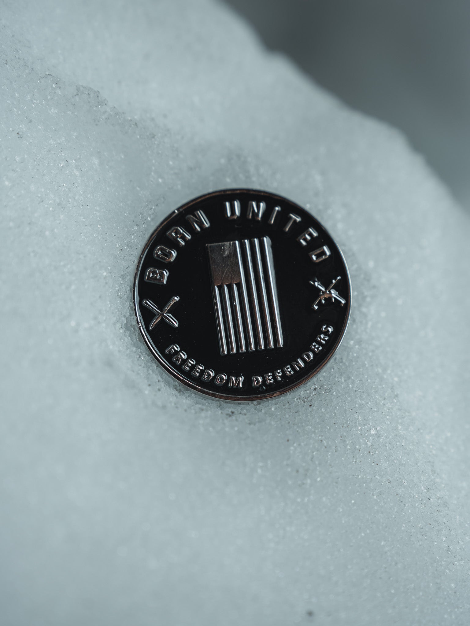 The Daddy’s Home Challenge Coin from Born United Private Label is a round metal piece on snow, showcasing vertical stripes and two crossed axes. The edges display BORN UNITED at the top and FREEDOM DEFENDERS at the bottom, symbolizing unity and resilience.
