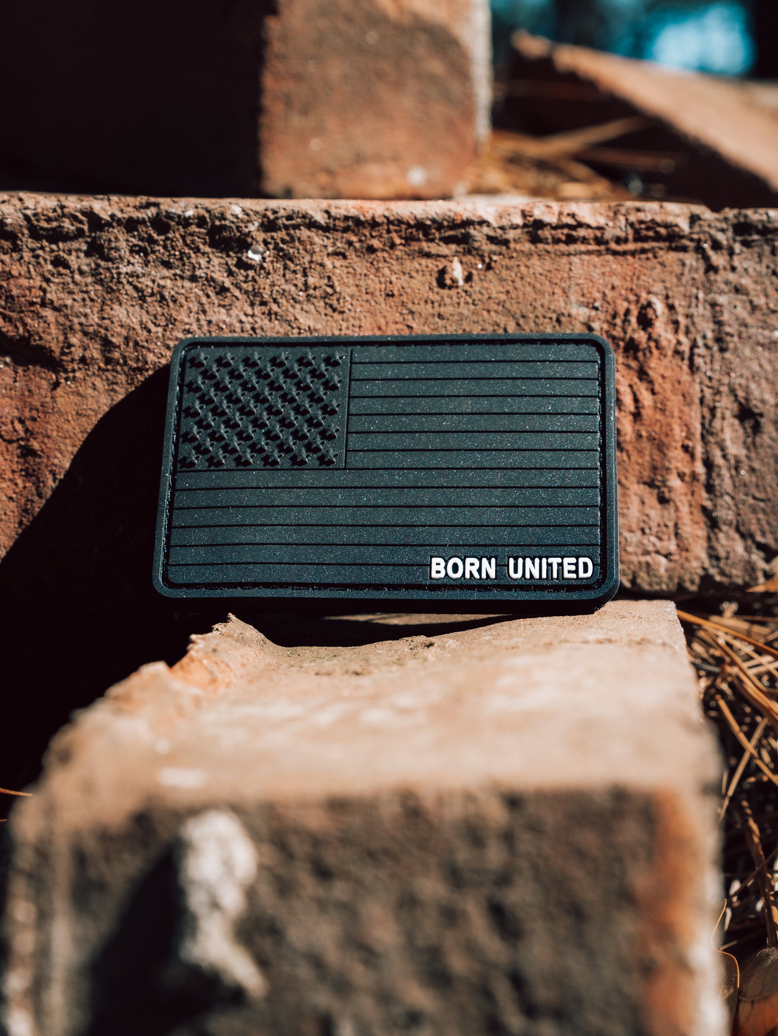 The Born United Private Labels American Flag PVC Patch - Blackout, displaying a U.S. flag and BORN UNITED, rests on sunlit, weathered bricks and includes a hook & loop backing.