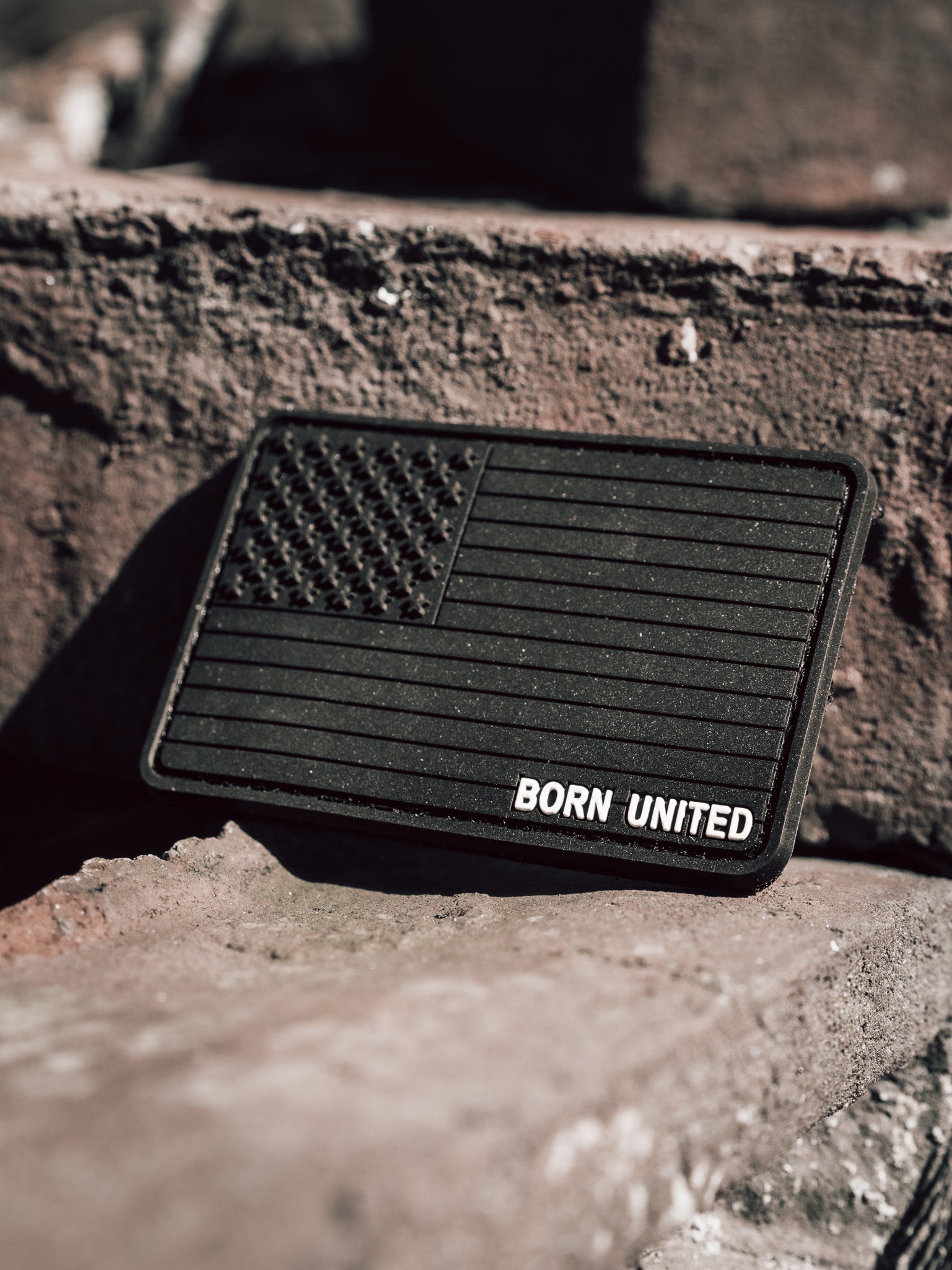 The American Flag PVC Patch - Blackout by Born United Private Label features a monochrome flag with BORN UNITED below, set against a rough brick surface. Angled sunlight highlights its contours, and the hook & loop backing allows for easy attachment.