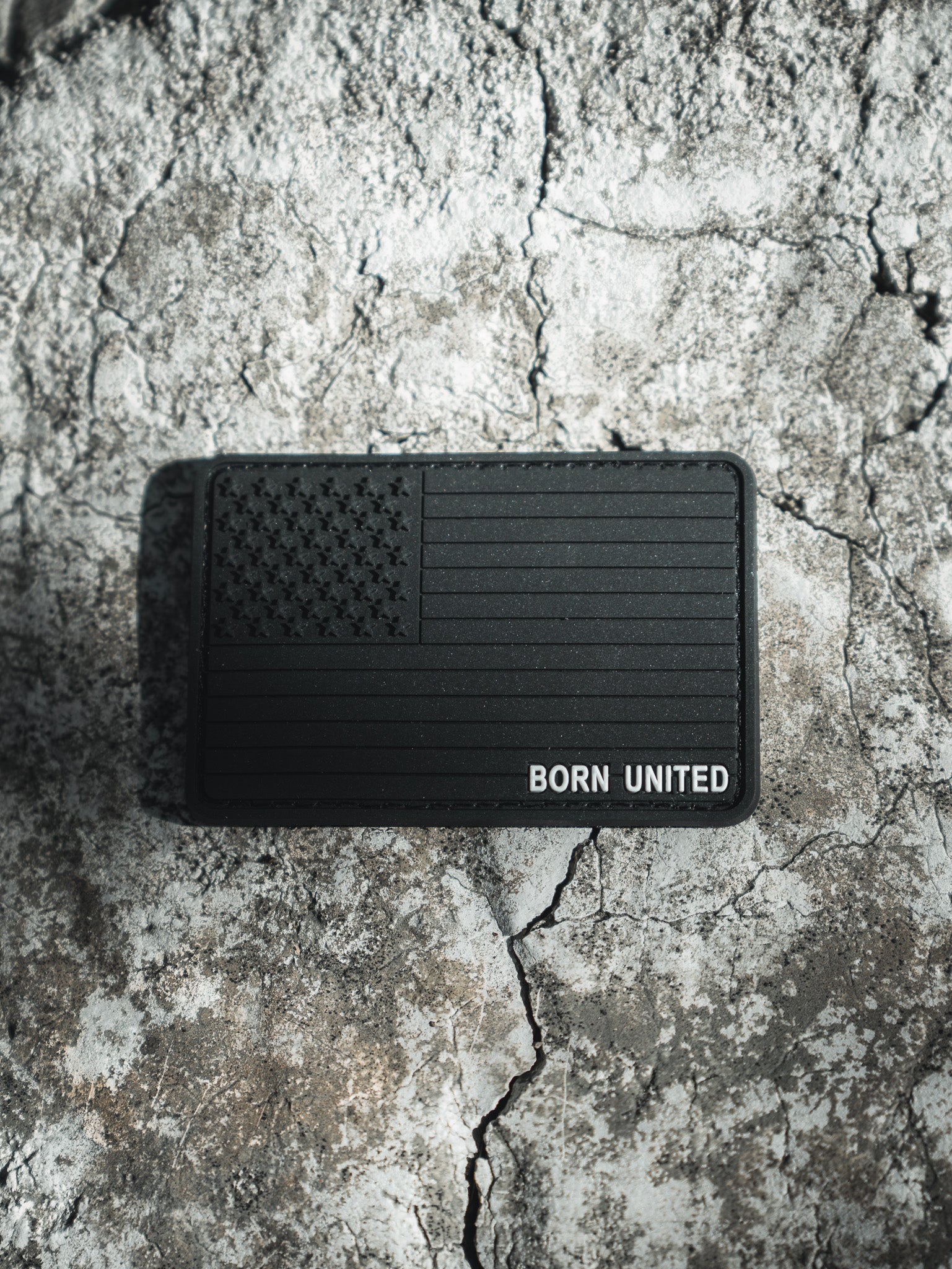 The Born United Private Labels American Flag PVC Patch - Blackout has a textured design featuring the black American flag with BORN UNITED, set against a rugged, stone-like surface. Its hook & loop backing ensures easy attachment, emphasizing its standout design against the rough background.