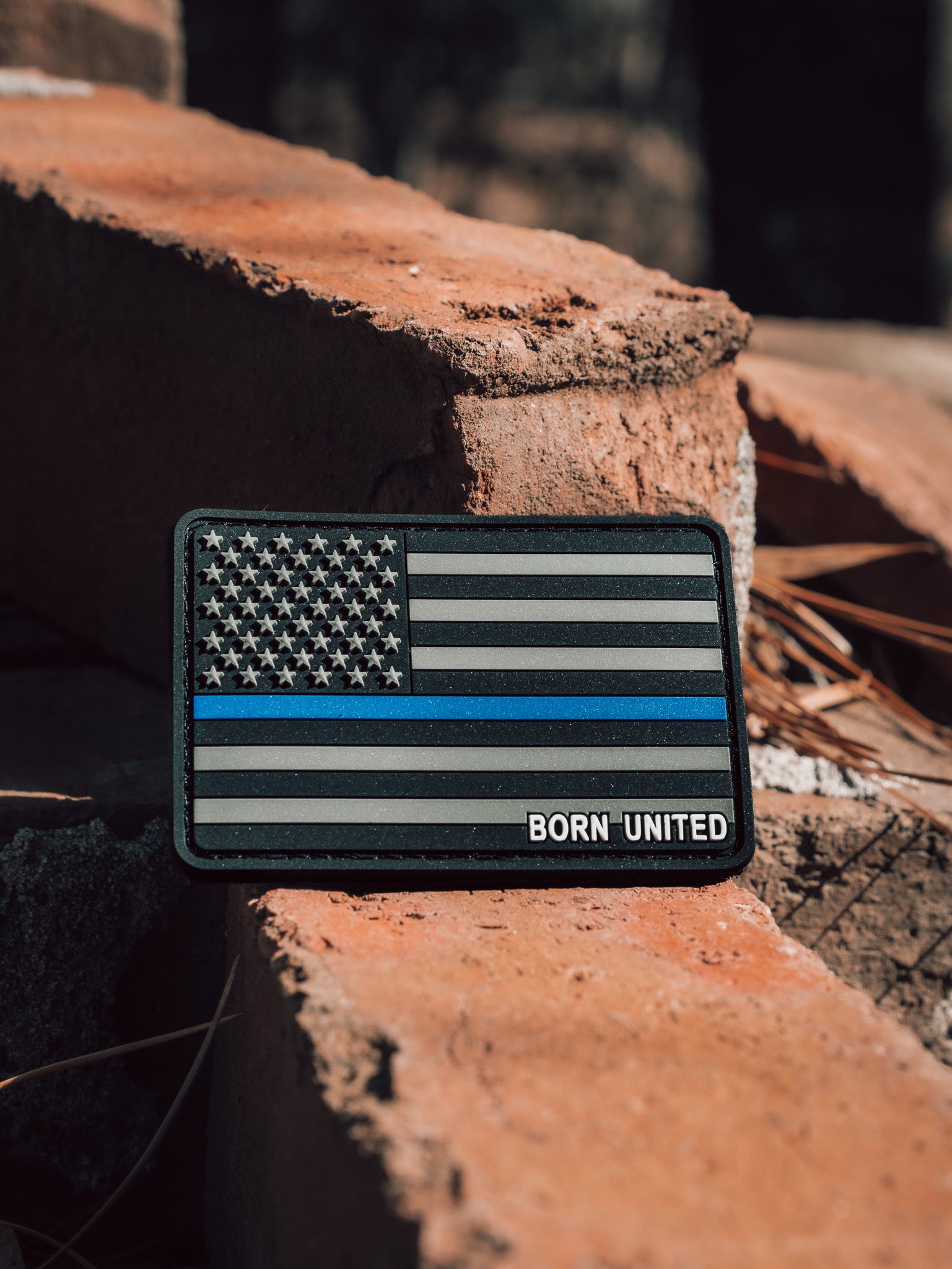 A durable PVC patch by Born United Private Label, featuring a monochrome American flag with a Thin Blue Line, rests on sunlit red bricks.