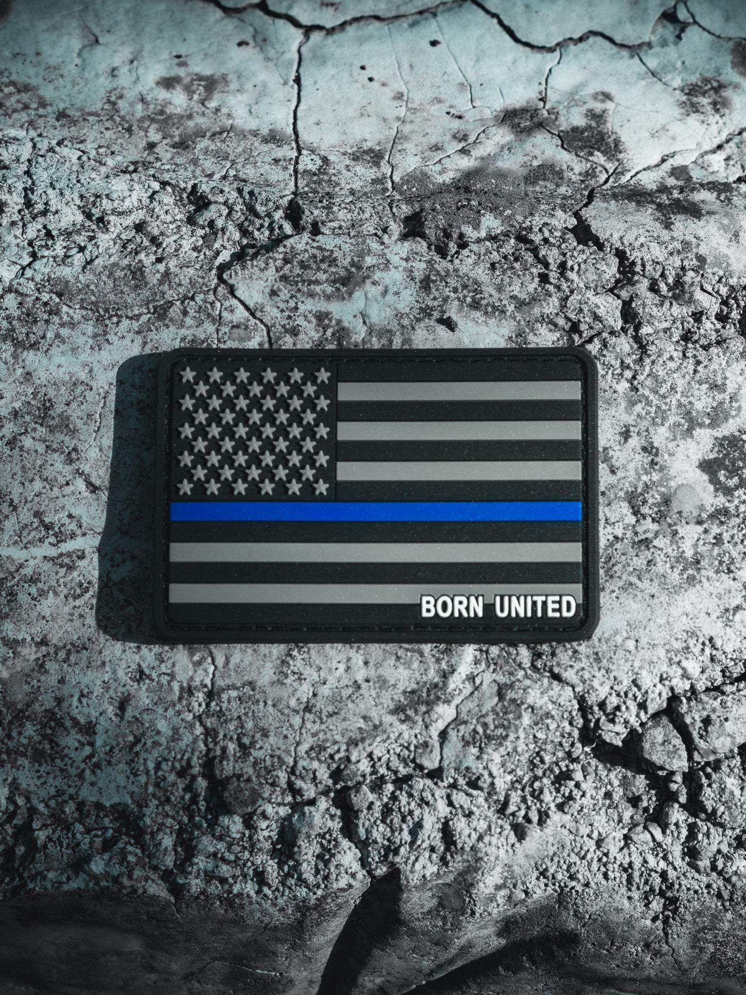 A resilient PVC patch named American Flag PVC Patch - TBL by Born United Private Label, features a grayscale flag with a bold blue stripe symbolizing law enforcement support, resting on cracked earth. Born United is elegantly inscribed in the lower right corner.
