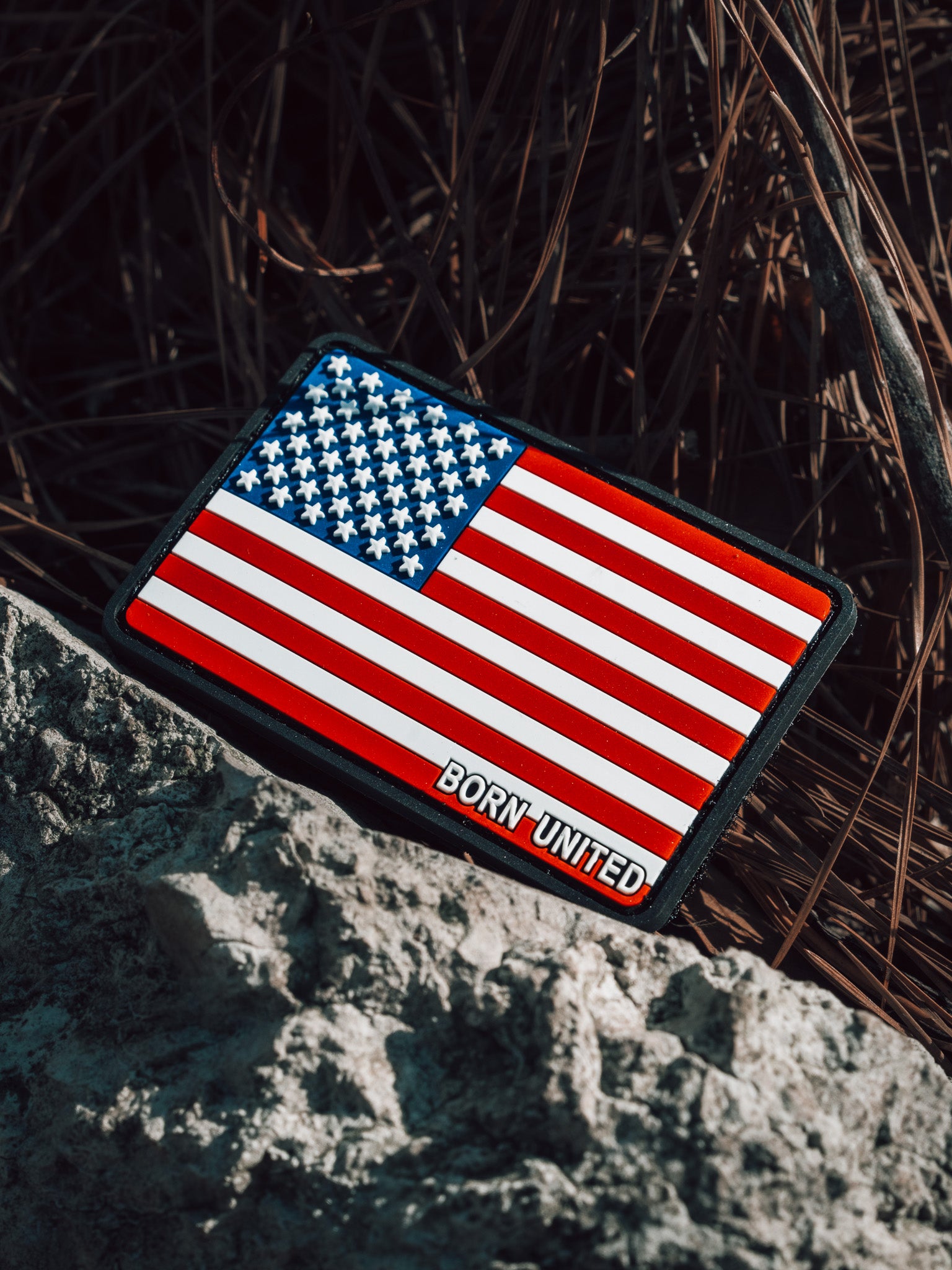 The American Flag PVC Patch by Born United Private Label lies on gray rocks against dried grass. Its hook & loop design is waterproof and washable, ensuring durability in any conditions.