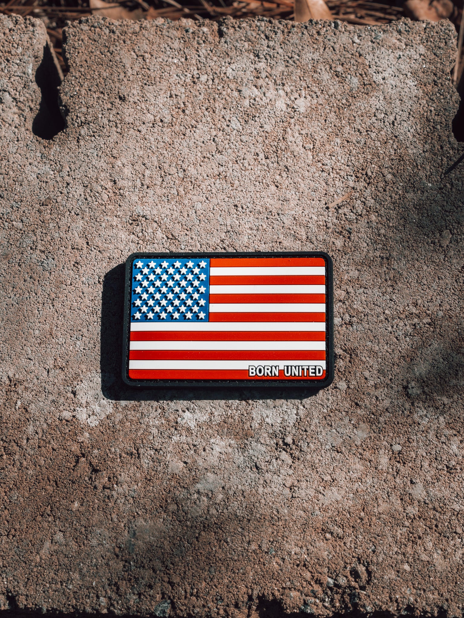 The American Flag PVC Patch by Born United Private Label rests on a rugged cement surface, with Born United boldly displayed. Its waterproof and washable, ensuring durability even in harsh conditions.