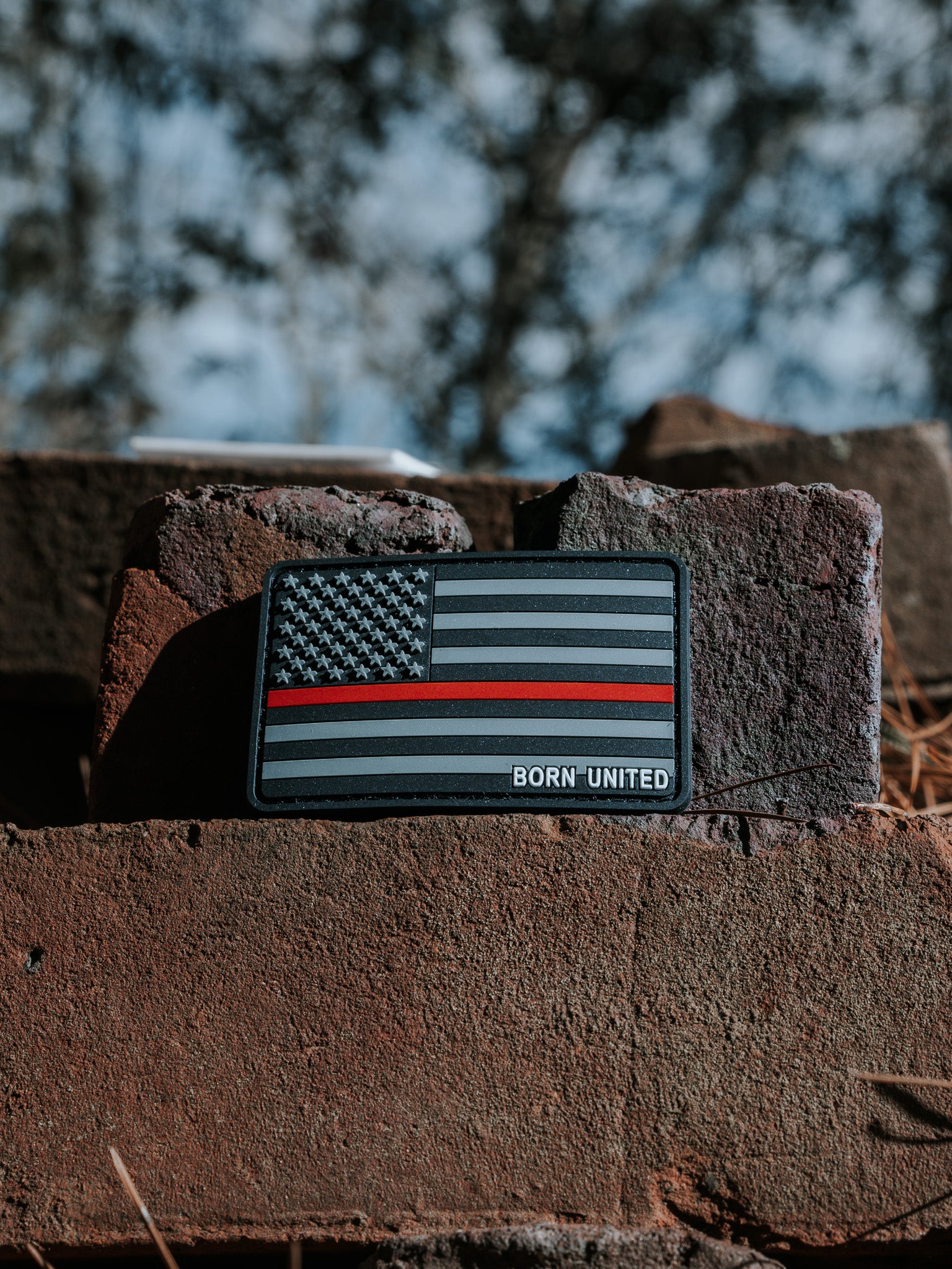 A Born United Private Label PVC patch, featuring a black and gray American flag with a red stripe symbolizing firefighter support, is showcased on a rough brick surface. The words BORN UNITED are visible against a blurred natural background.
