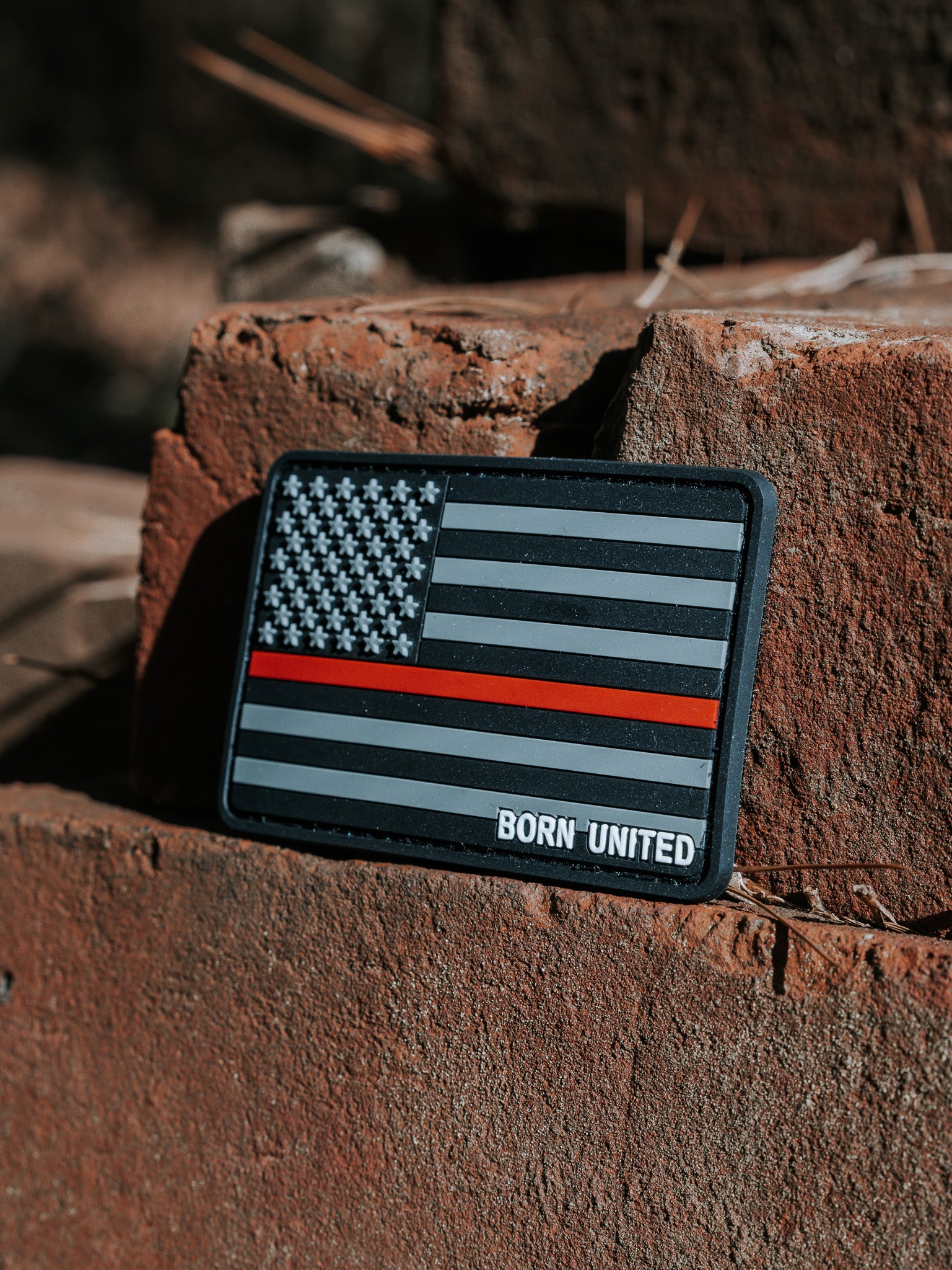 An American Flag PVC Patch - TRL by Born United Private Label, featuring a Thin Red Line and BORN UNITED text below, is displayed on weathered bricks outdoors with natural light casting complex shadows.