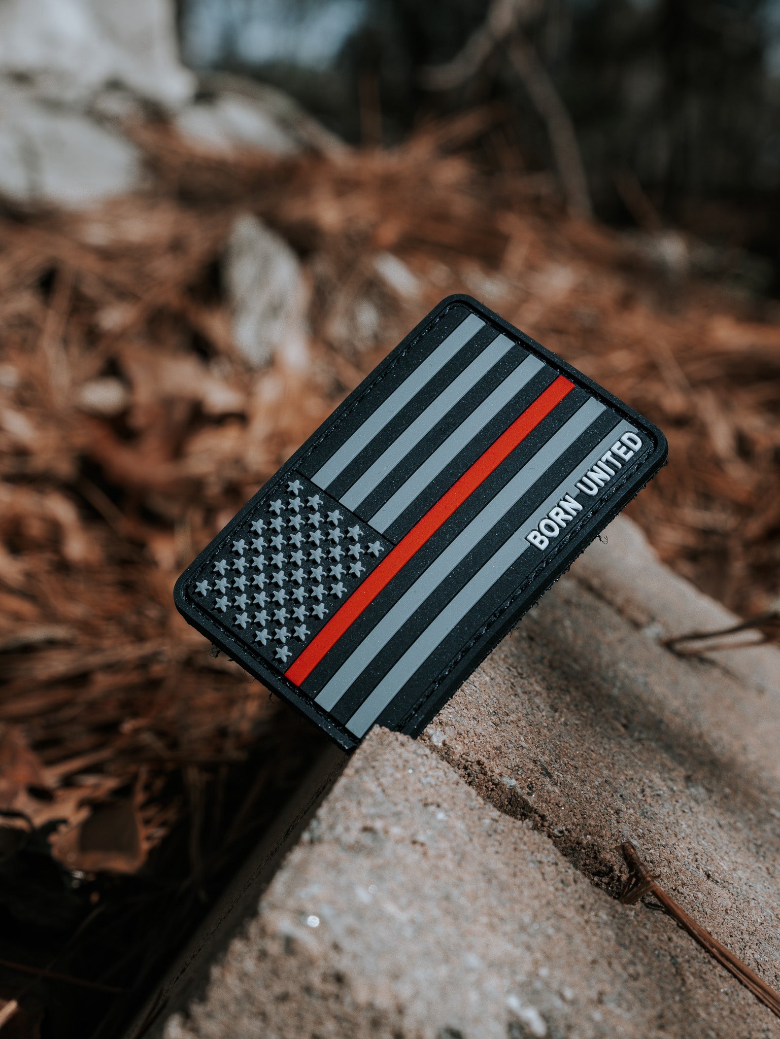 The American Flag PVC Patch - TRL by Born United Private Label features a black and gray Thin Red Line design, symbolizing firefighter support. It rests against a concrete block with dried leaves and pine needles in the background.
