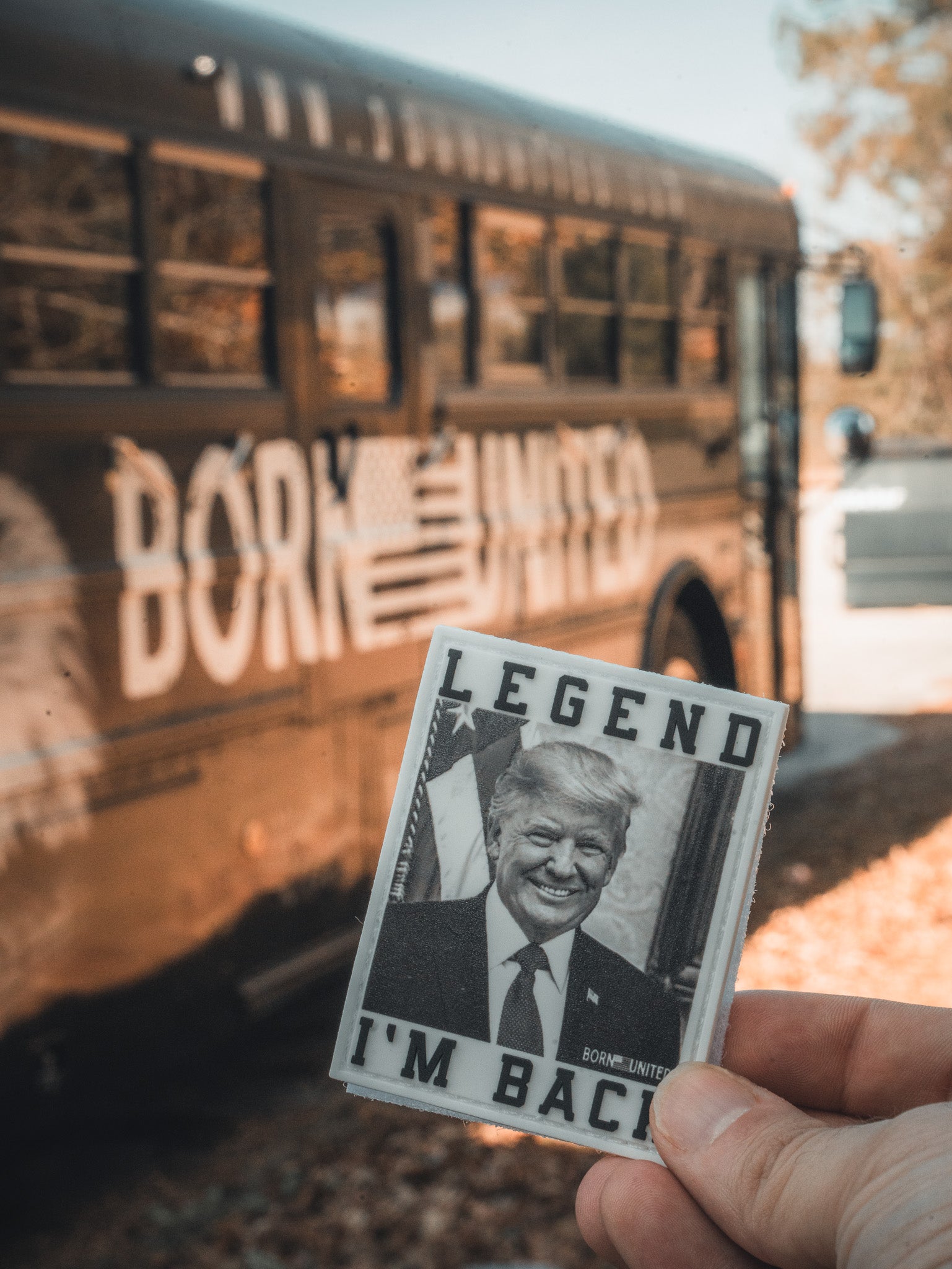 A hand holds an Im Back patch from Born United Private Label featuring a photo of a man in a suit. A dark-colored bus with Born United and an American flag design is in the background, with trees and sunlight reminiscent of Trump 2024s historical win.