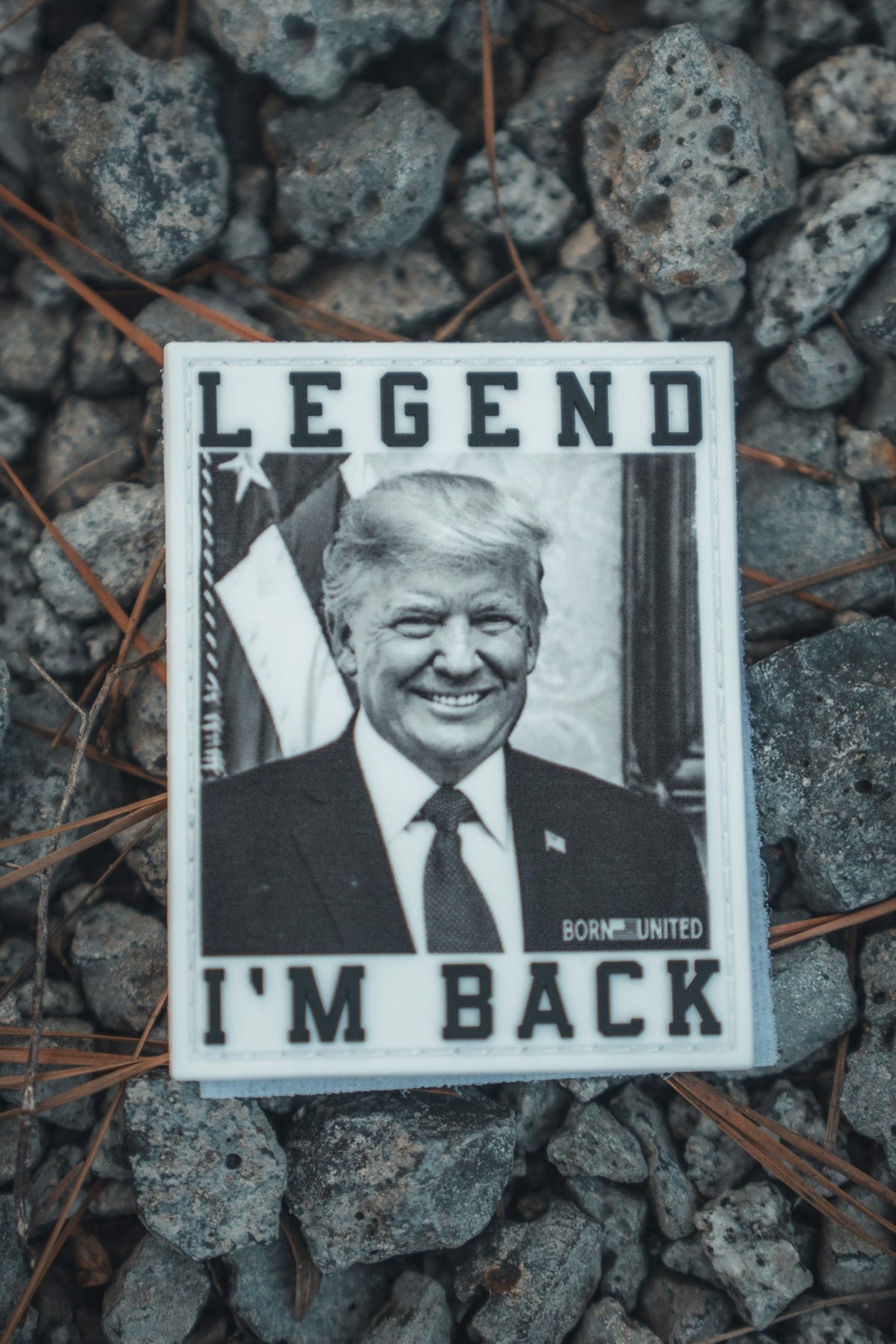 A photo of the Born United Private Label “I’m Back” PVC Patch, showing a black-and-white suited man smiling with “LEGEND” on top and “I’M BACK” below, lying on gravel with pine needles—a nod to President Trump 2024.