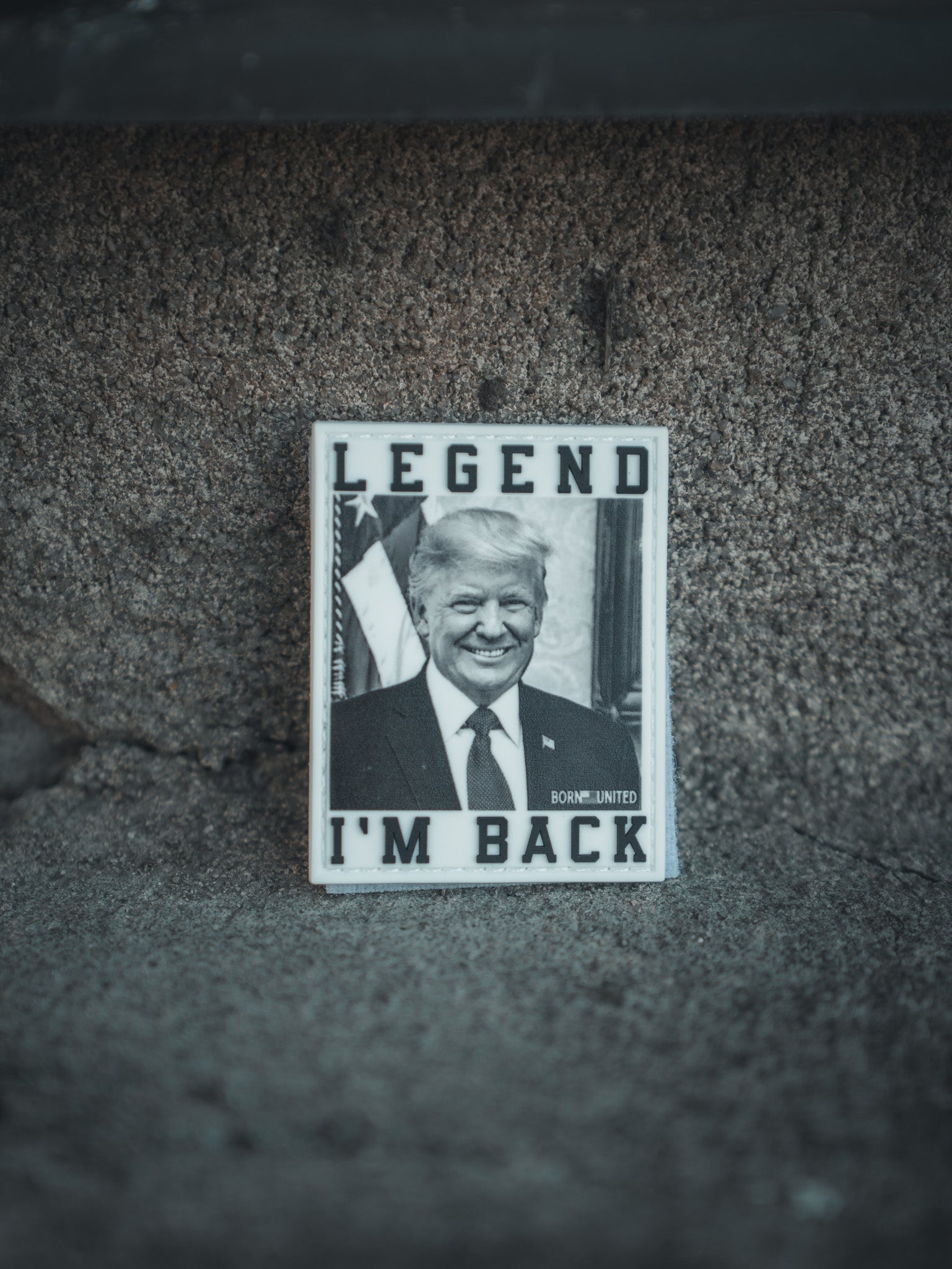 A photograph shows a man on a textured surface wearing a suit and tie with an American flag backdrop. The top reads LEGEND, and the bottom says IM BACK. A proud presidential patch hints at Trump 2024. Available as the “I’m Back Patch” from Born United Private Label.