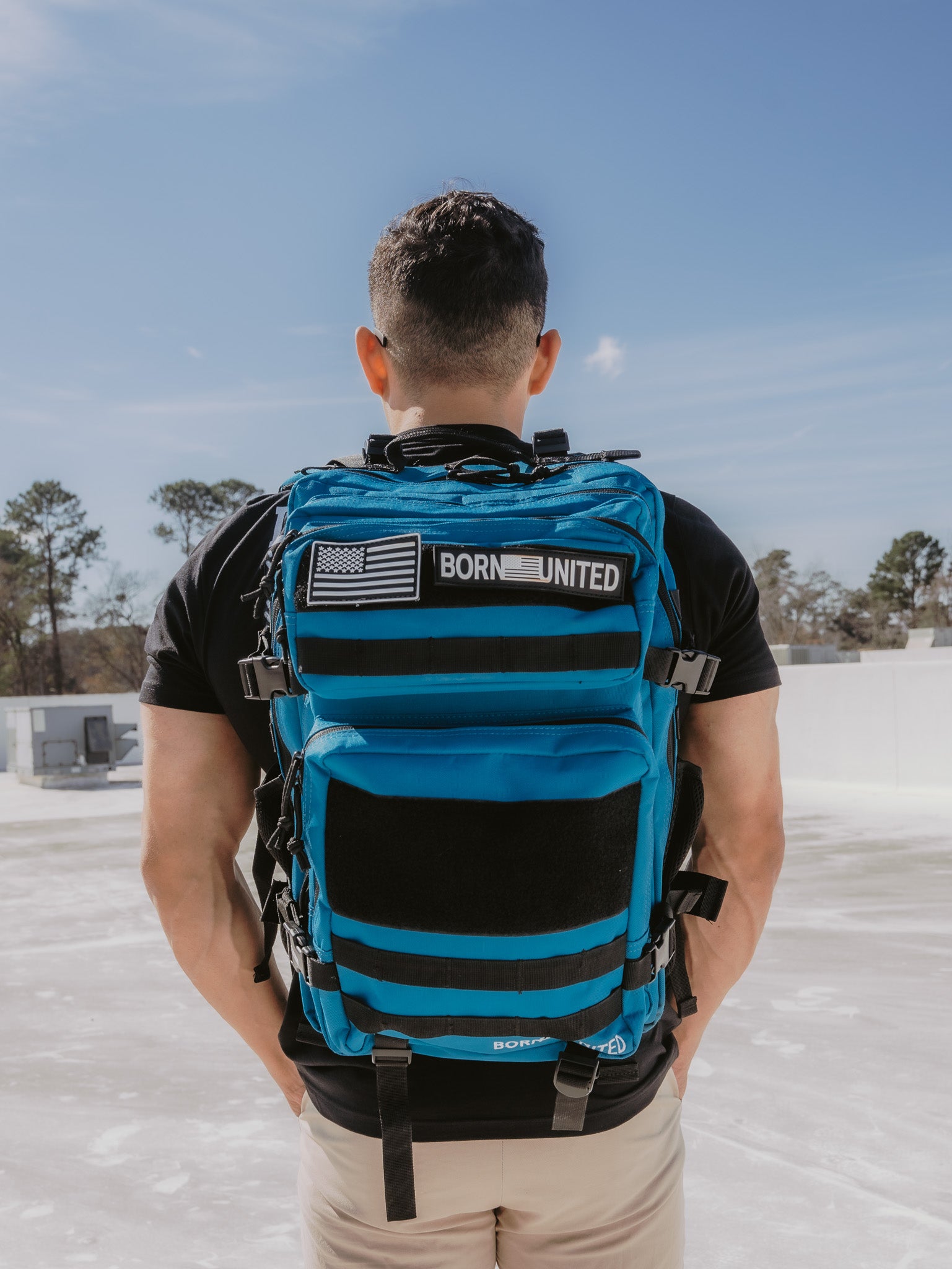 A person with short hair stands outside, back turned, wearing a black shirt and beige pants. They carry the 45L Electric Blue Premium Tac-Pac by Born United Private Label, featuring black straps and an American flag patch. The sky is clear, with trees visible in the background.