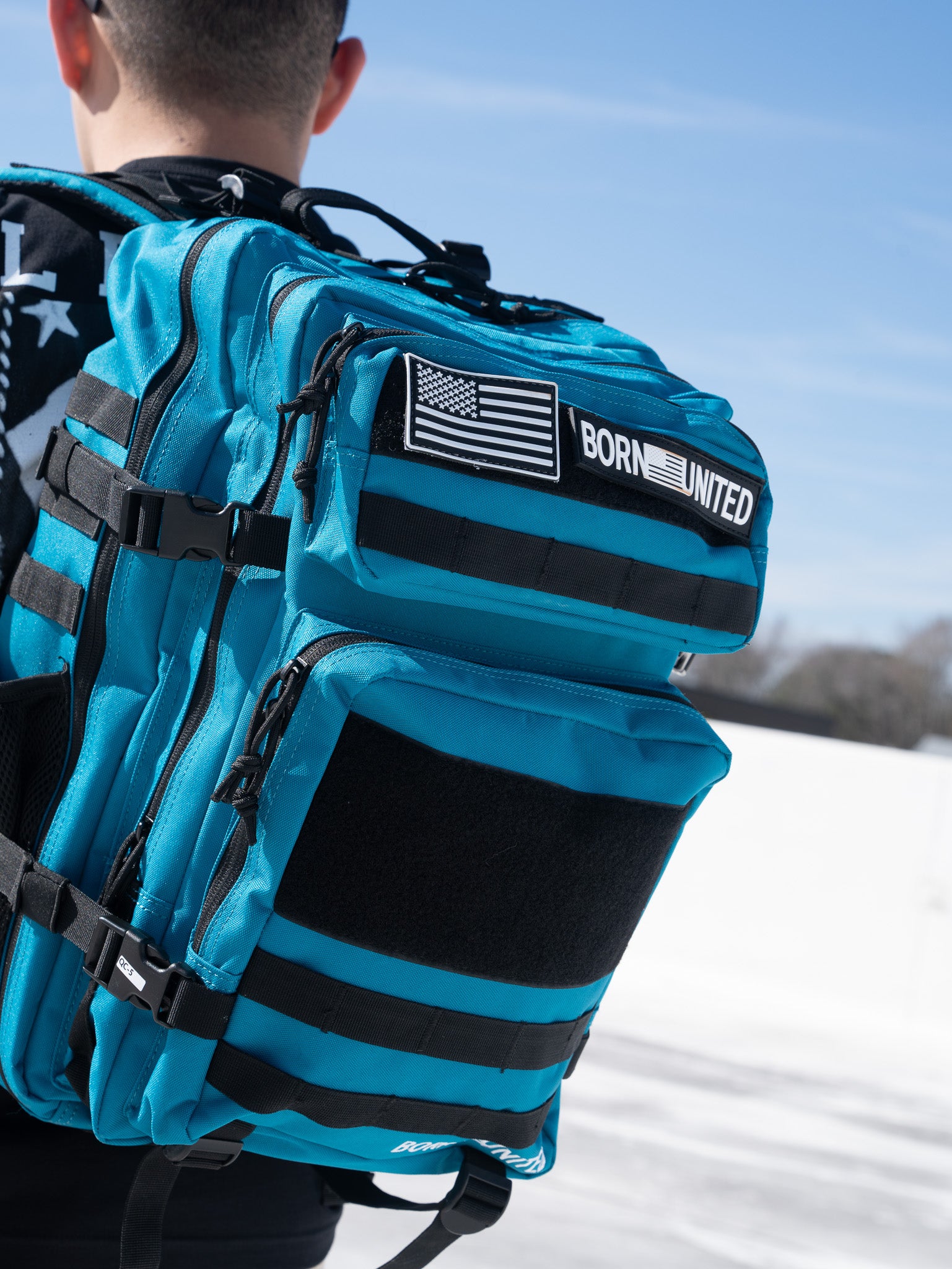 A person with short hair wears a large Electric Blue Premium Tac-Pac - 45L by Born United Private Label, featuring black straps, an American flag patch, and a BORN UNITED patch. The sky is bright and clear with trees visible in the distance.
