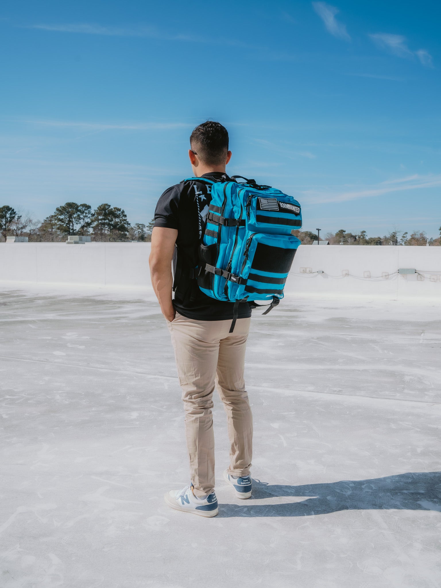 With their back to the camera, a person is outdoors in a black t-shirt, beige pants, and white sneakers. They carry a Born United Private Label Electric Blue Premium Tac-Pac - 45L backpack that contrasts vividly against the clear sky while standing on a paved surface with distant trees.