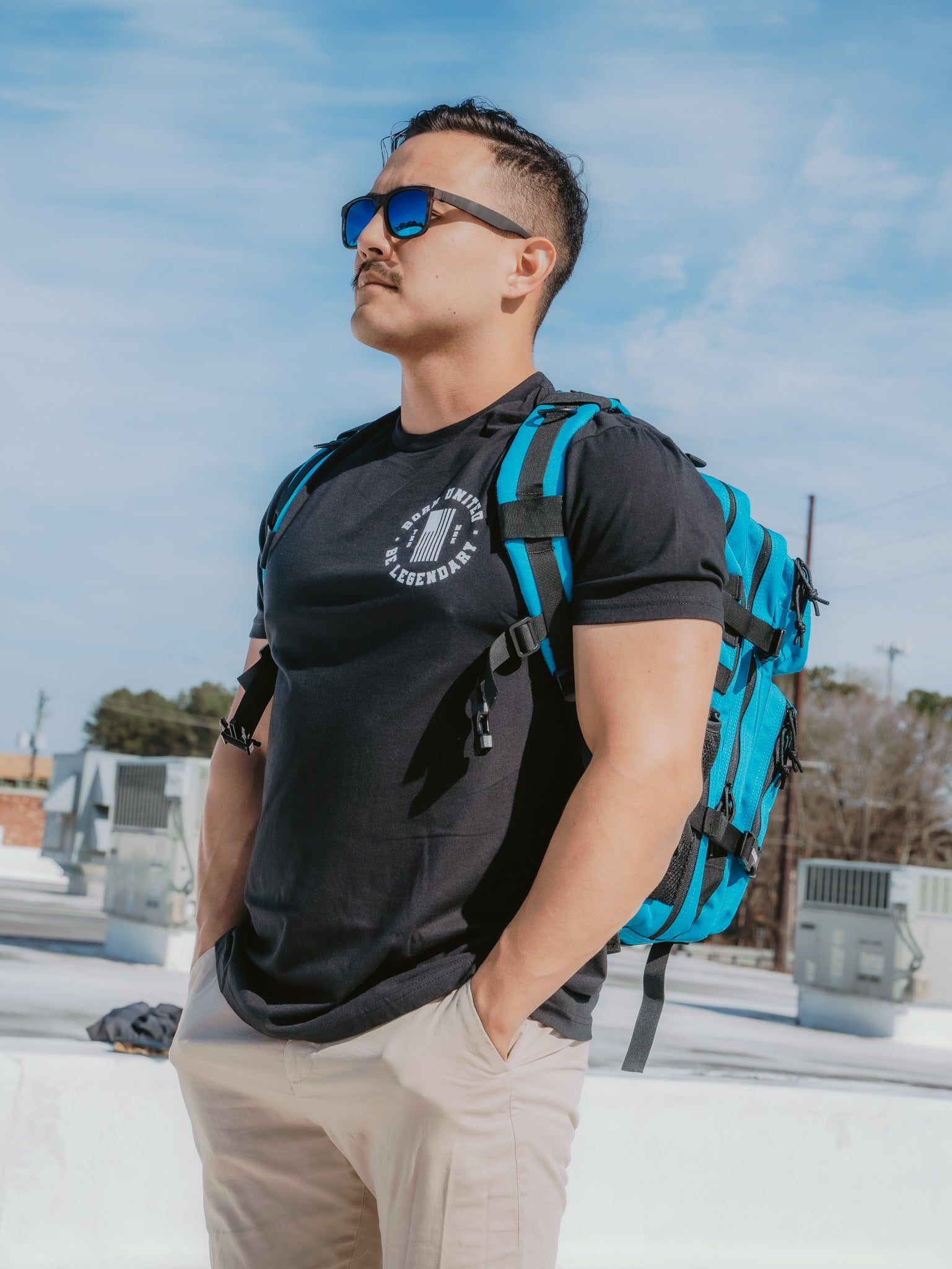 A man with short hair, wearing electric sunglasses and a black T-shirt, pairs his look with beige pants and the Electric Blue Premium Tac-Pac - 45L by Born United Private Label. He stands outdoors confidently with hands in pockets, as the sky and buildings set the scene behind him.
