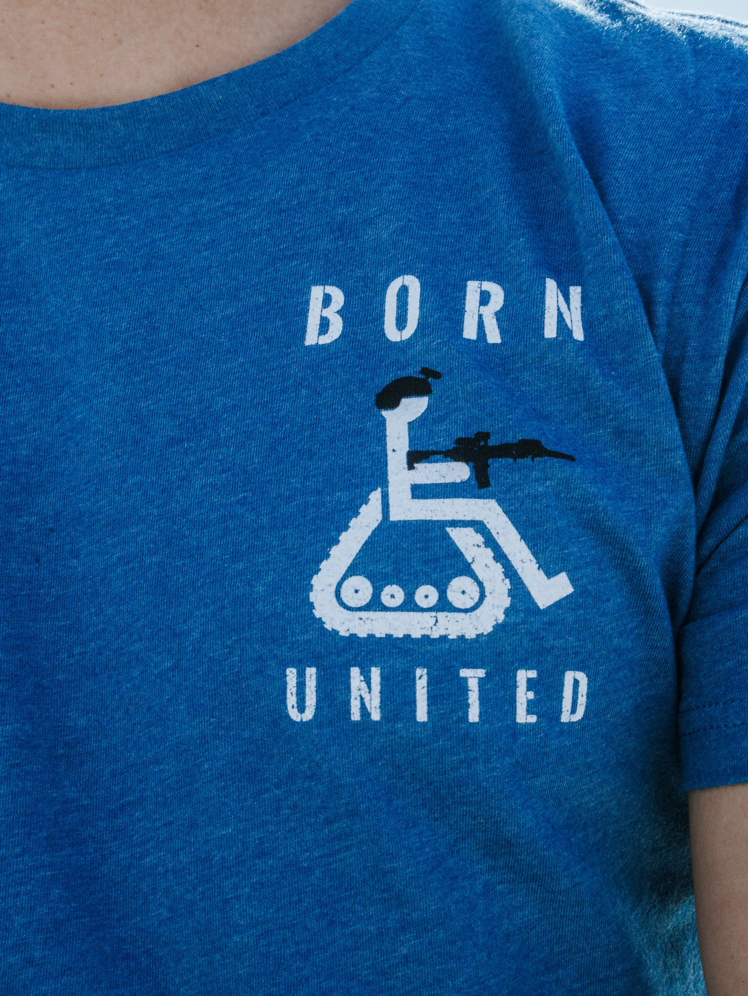 A person is wearing the Disabled Vet - Blue t-shirt by Born United Private Label, showcasing a disgruntled wheelchair symbol with tank tracks and a figure with a rifle, along with the words BORN and UNITED boldly printed above and below.