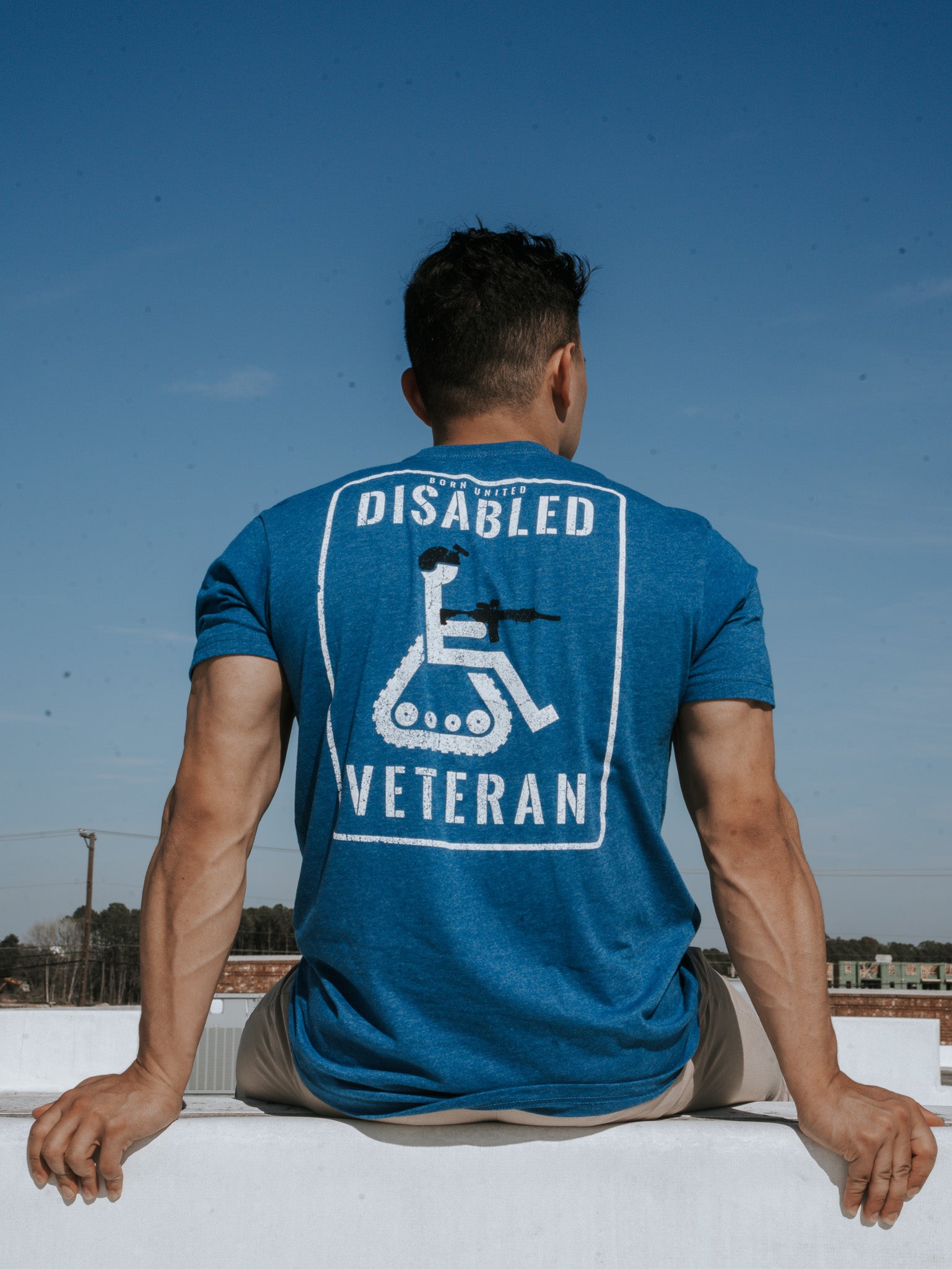 A disgruntled person sits on a ledge against a clear blue sky, wearing the Disabled Vet - Blue T-shirt by Born United Private Label, featuring a graphic of a wheelchair and the text Disabled Veteran.