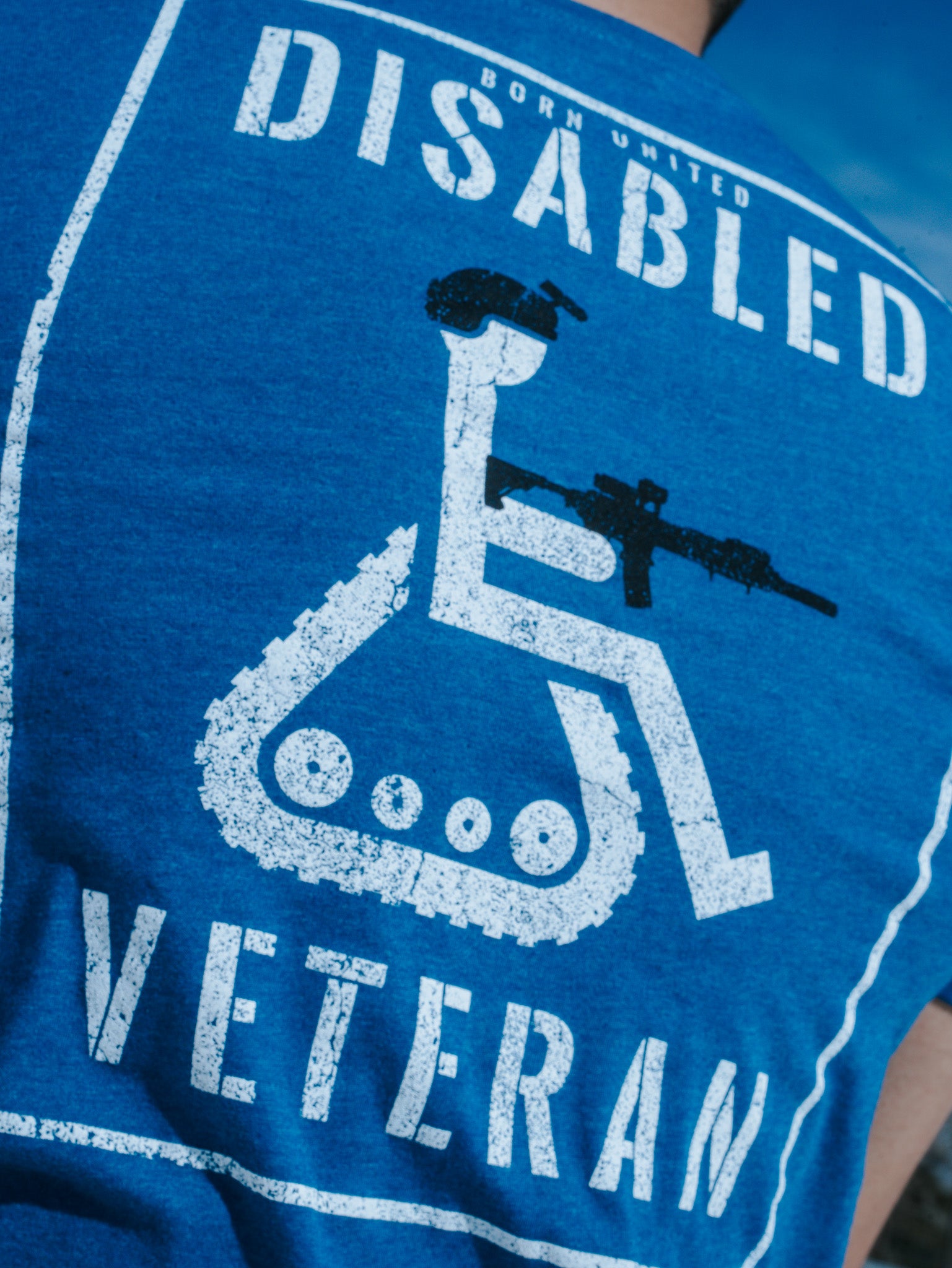 Back view of the Born United Private Label Disabled Vet - Blue T-shirt, featuring a wheelchair symbol with a helmet and rifle, along with Disabled Veteran text. The wearers disgruntled expression complements the outdoor setting against a tranquil blue sky.