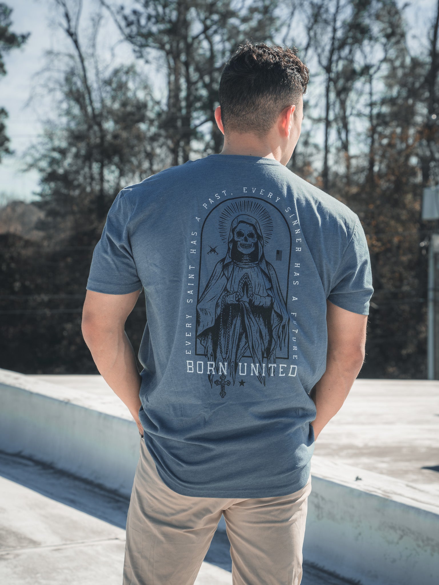 A man outdoors on a rooftop faces away, wearing the Saints And Sinners Tee- Blue by Born United Private Label. The shirt showcases a hooded figure with the text Every Saint Has a Past, Every Sinner Has a Future.