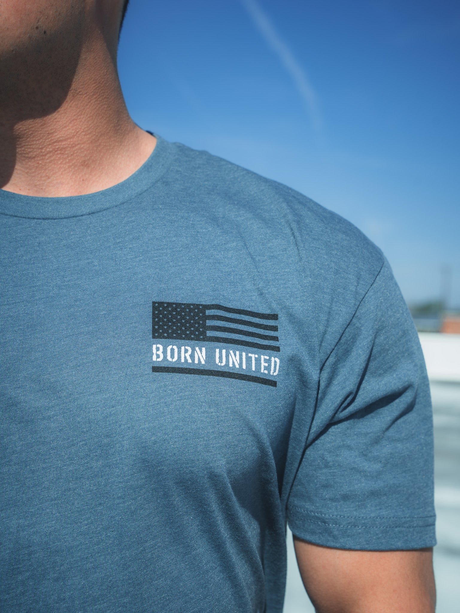 A person wearing the Saints And Sinners Tee- Blue by Born United Private Label, featuring a black and white U.S. flag with BORN UNITED on the chest, stands against a clear blue sky, seemingly pondering the future. Only their upper torso is visible.