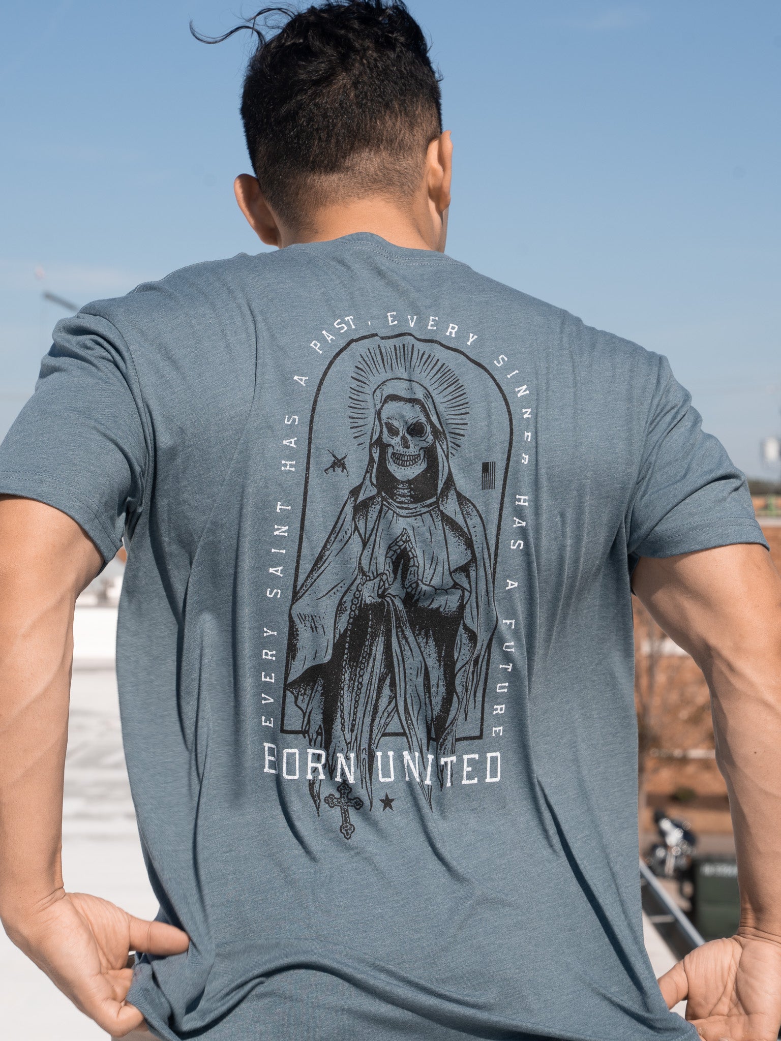 A person is wearing the Saints And Sinners Tee- Blue by Born United Private Label, featuring a skeletal saint in a robe with a halo and prayer hands. The text reads, Every saint has a past, every sinner has a future, with Born United below.