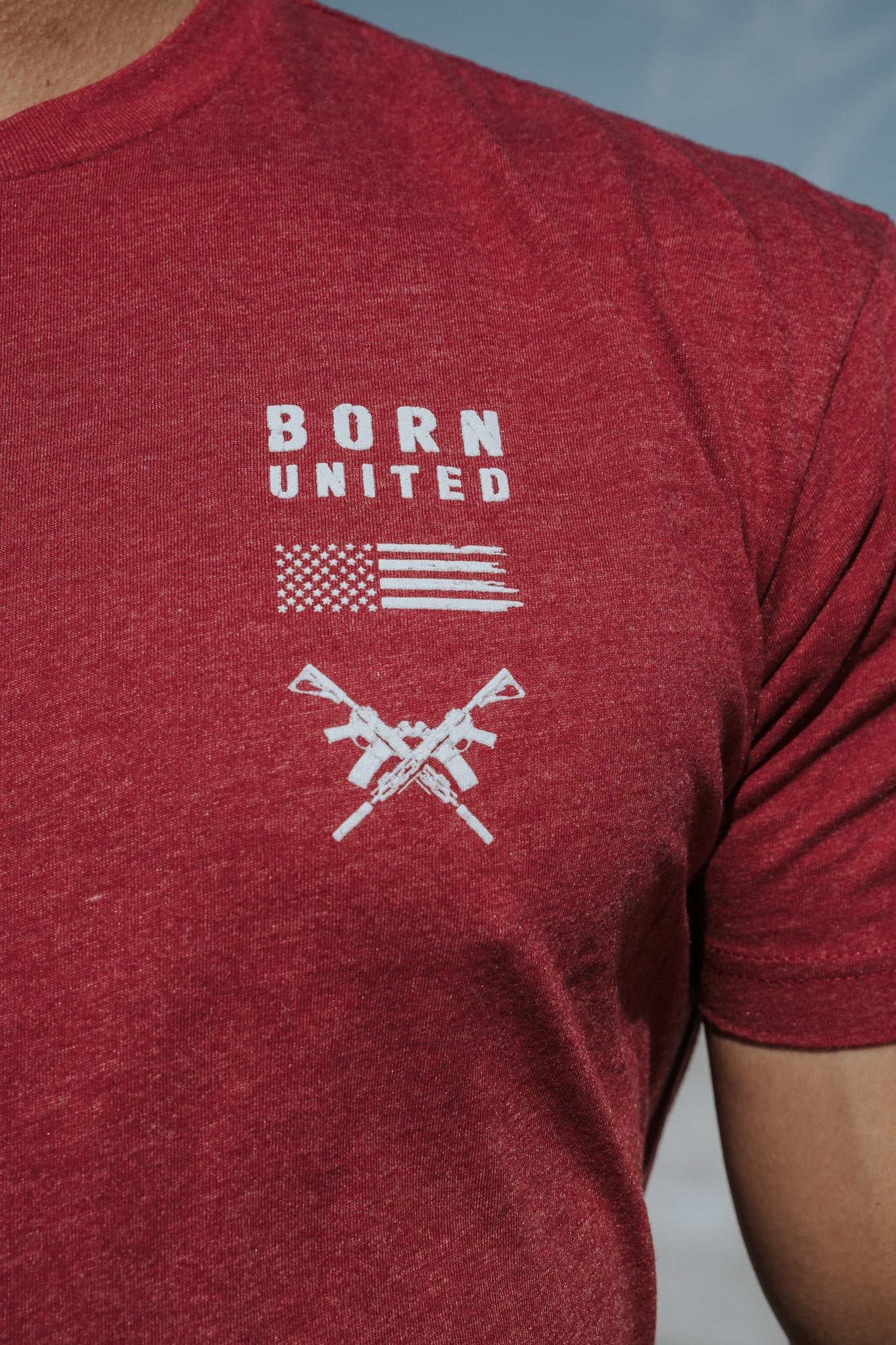 A person wears the R.E.D. Friday T-shirt by Born United Private Label, featuring BORN UNITED text above a stylized American flag and crossed rifles, celebrating patriotism and honoring brave soldiers against a clear blue sky backdrop.