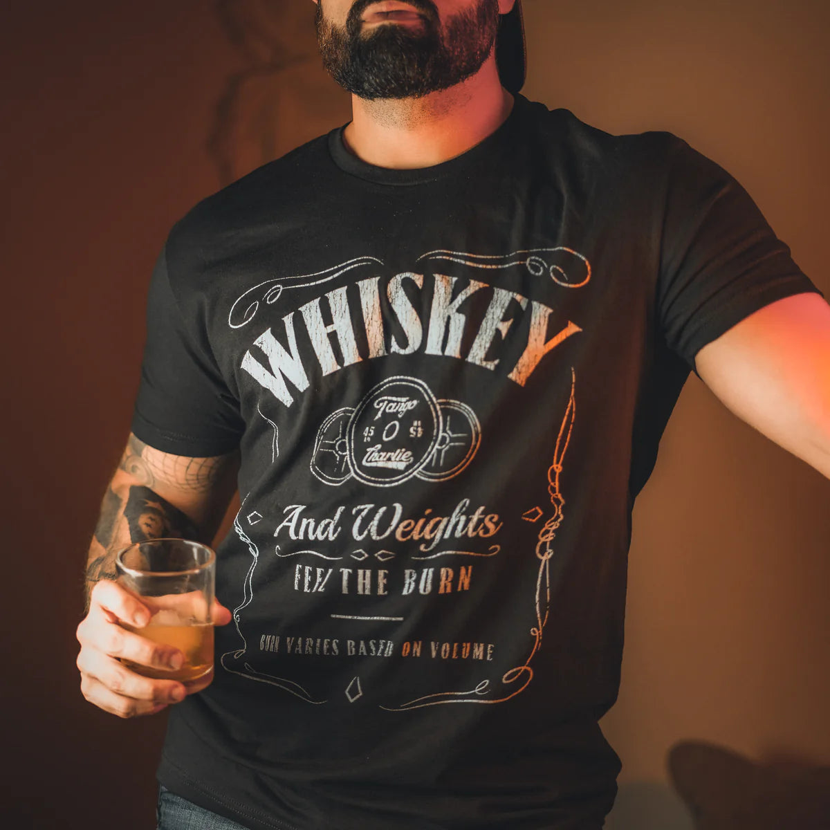 A bearded man in a black cap and Tango Charlie Apparels Whiskey Feel The Burn tee, which reads, WHISKEY And Weights, Feel the Burn, holds a whiskey glass. Tattoos cover his right forearm under warm lighting that sets an intimate mood.