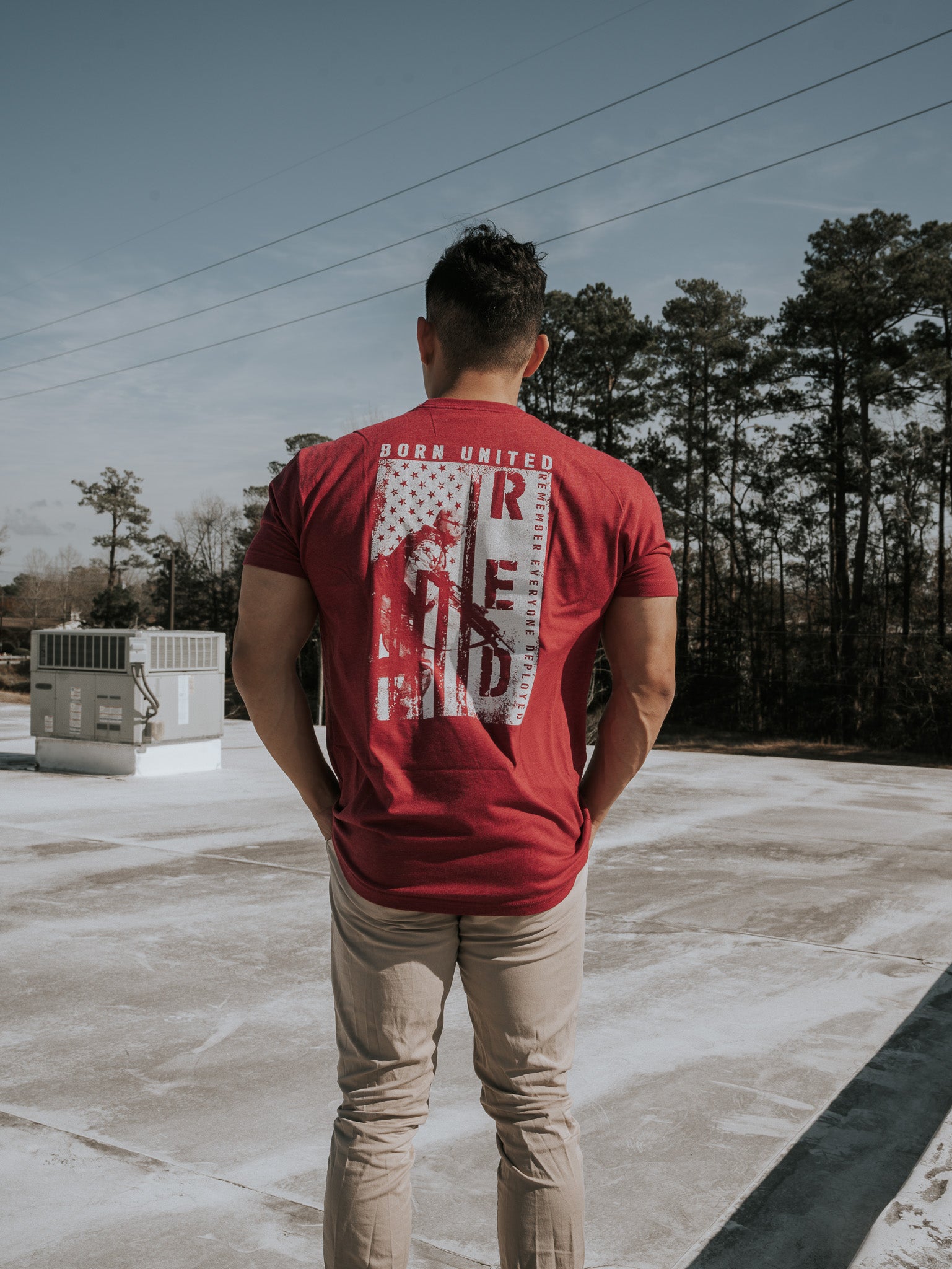 A man stands on a rooftop wearing a R.E.D. Friday shirt by Born United Private Label, facing away under a clear sky surrounded by trees, seemingly contemplating the freedoms protected by brave soldiers as power lines stretch across the horizon.