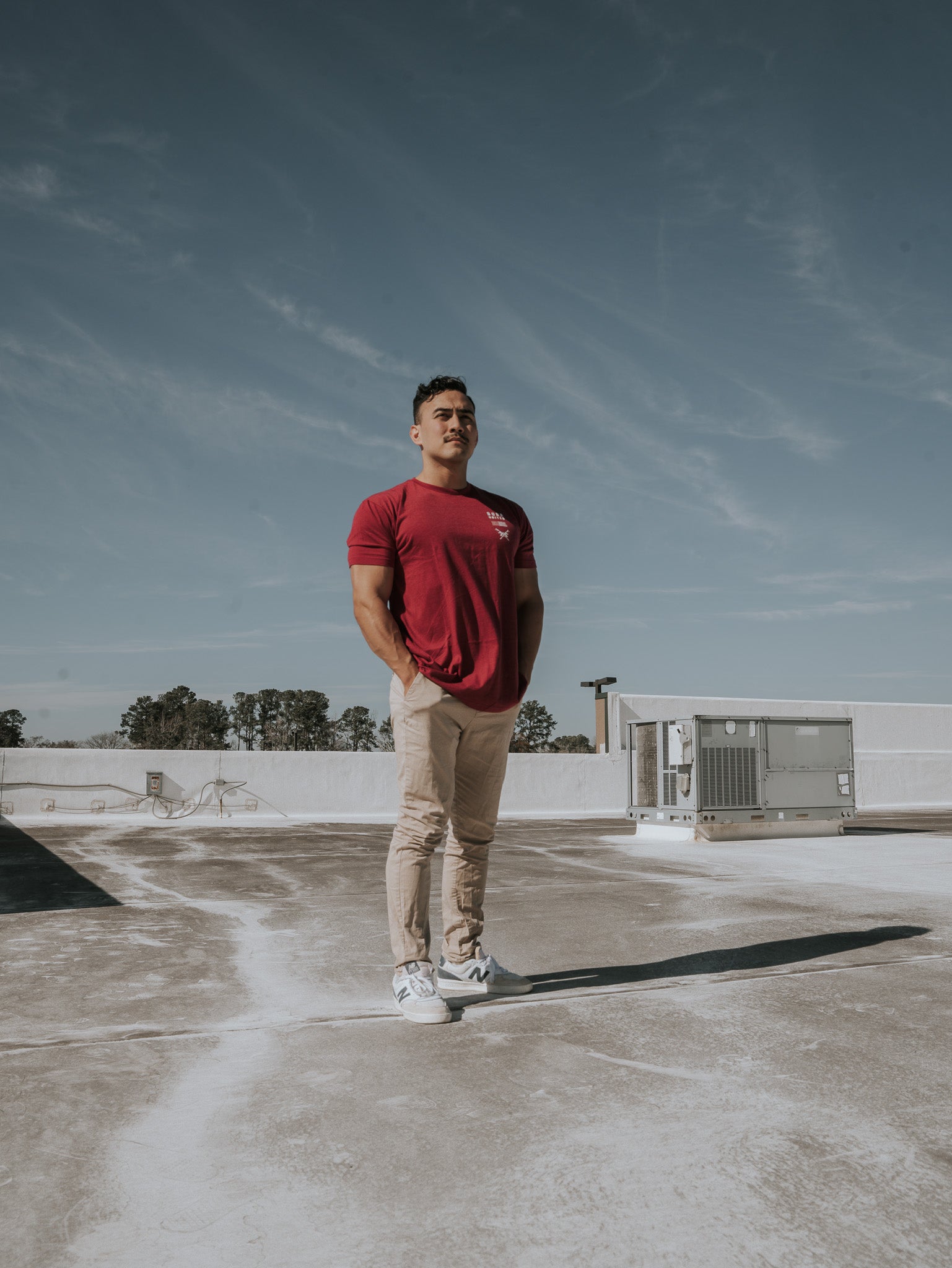A man in a Born United Private Label R.E.D. Friday T-shirt and beige pants stands on a concrete rooftop, hands in pockets under a clear blue sky. Air conditioning units stand like sentinels behind him, embodying the quiet patriotism reflected in everyday life.
