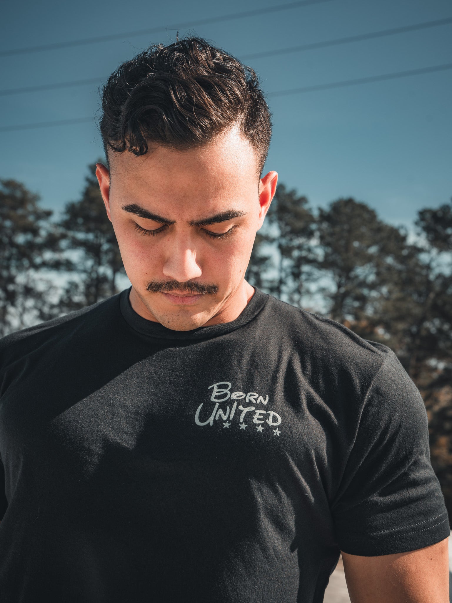 A man with short hair and a mustache wears the Triggered - Black Tee by Born United Private Label, looking downward. He stands outdoors against trees and a clear blue sky, embodying a fully strapped patriot vibe.