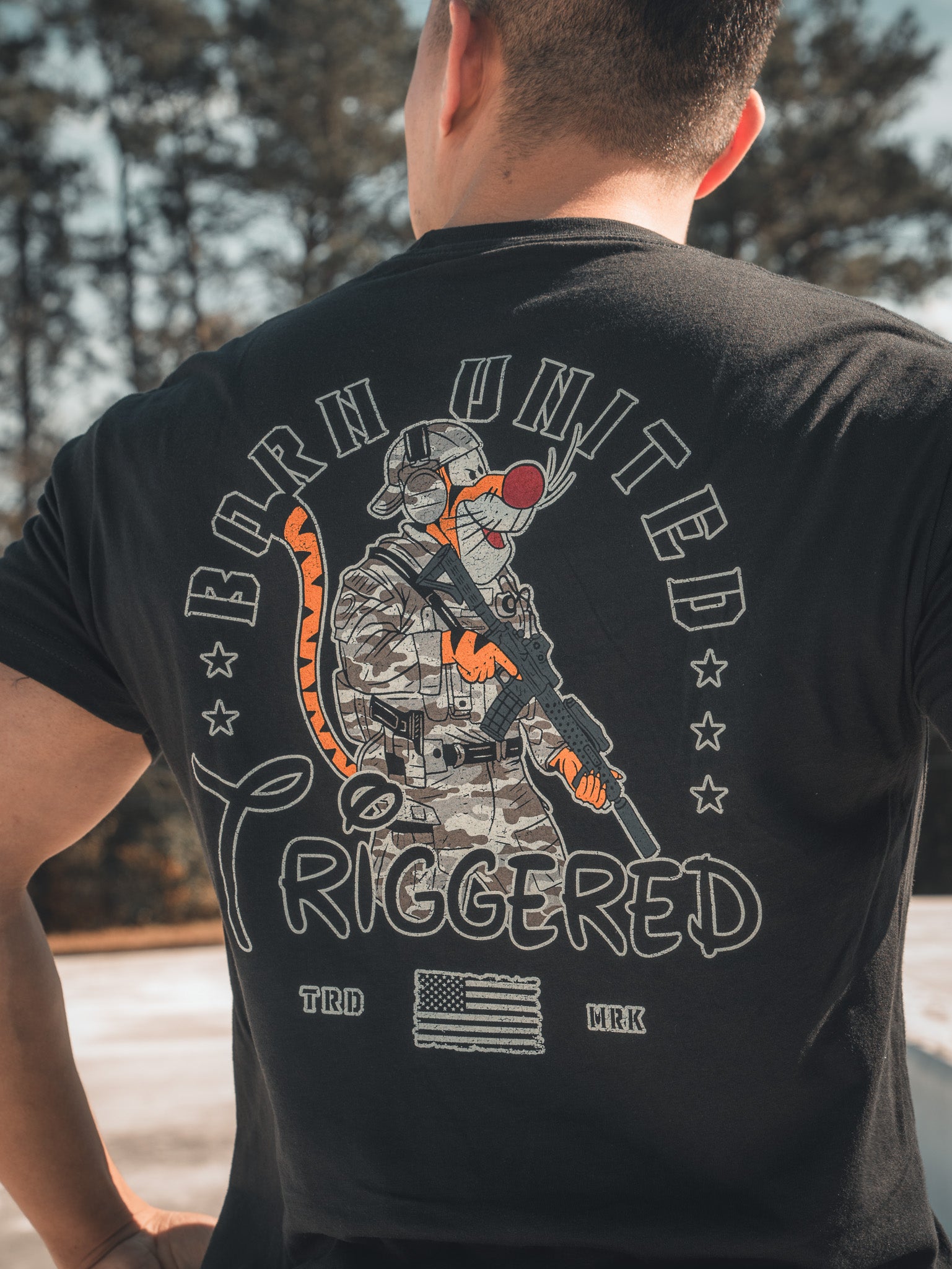 A fully strapped patriot sports the Triggered - Black Tee from Born United Private Label, featuring a cartoon soldier mouse in camouflage and sunglasses. The shirt reads Born United Triggered with a small American flag, against a backdrop of trees and snow for an American makeover.