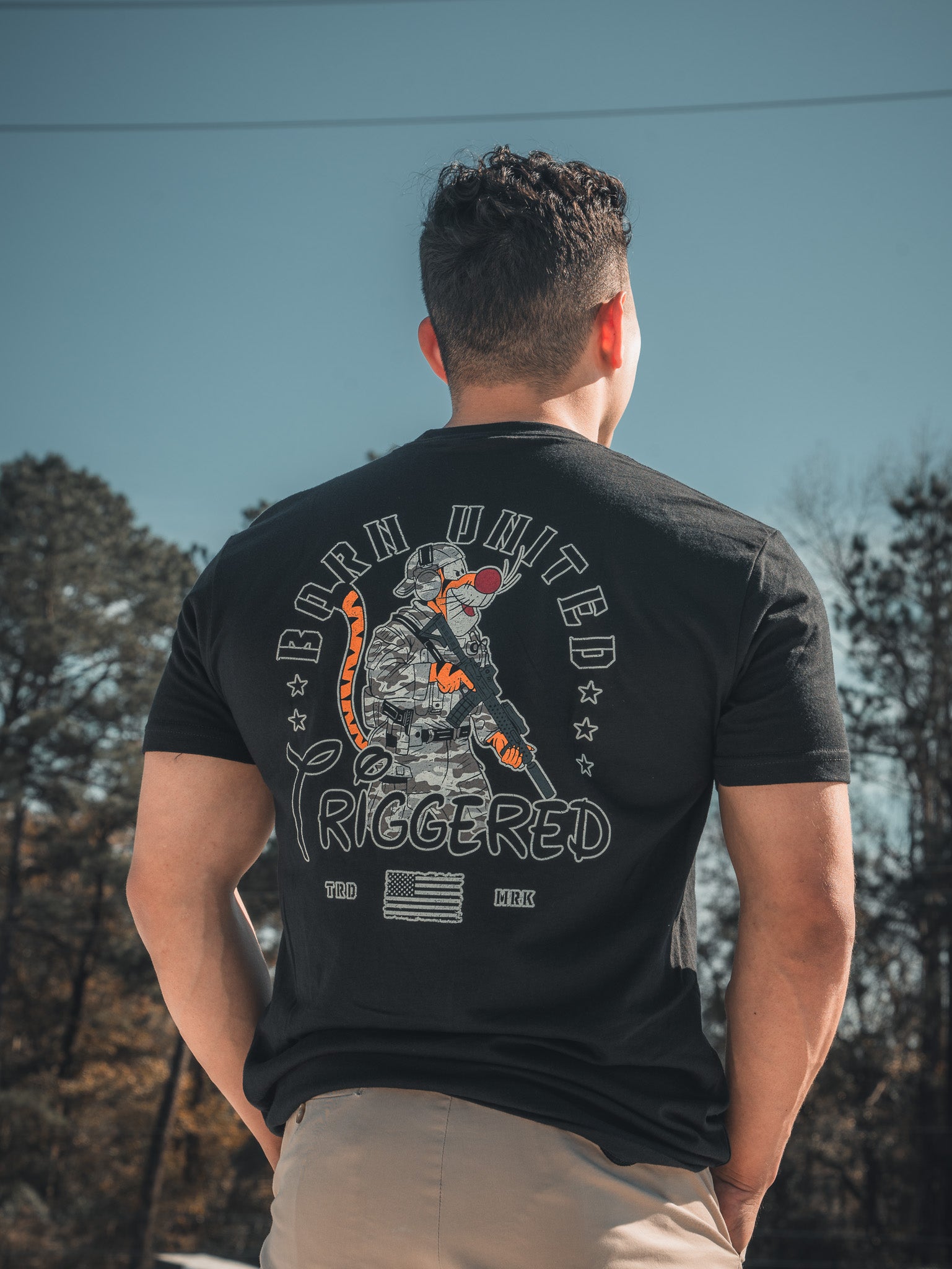A man stands amidst trees and a clear blue sky, wearing the Triggered - Black Tee by Born United Private Label. The t-shirts back features a military-themed graphic with Born United Triggered, showing an astronaut holding a rifle.