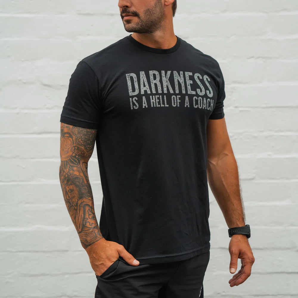A tattooed man stands against a light wall, looking to the side with one hand in his pocket, wearing the Tango Charlie Apparel Darkness Is A Hell Of A Coach T-Shirt in black, made from perfect stretch ring-spun cotton.