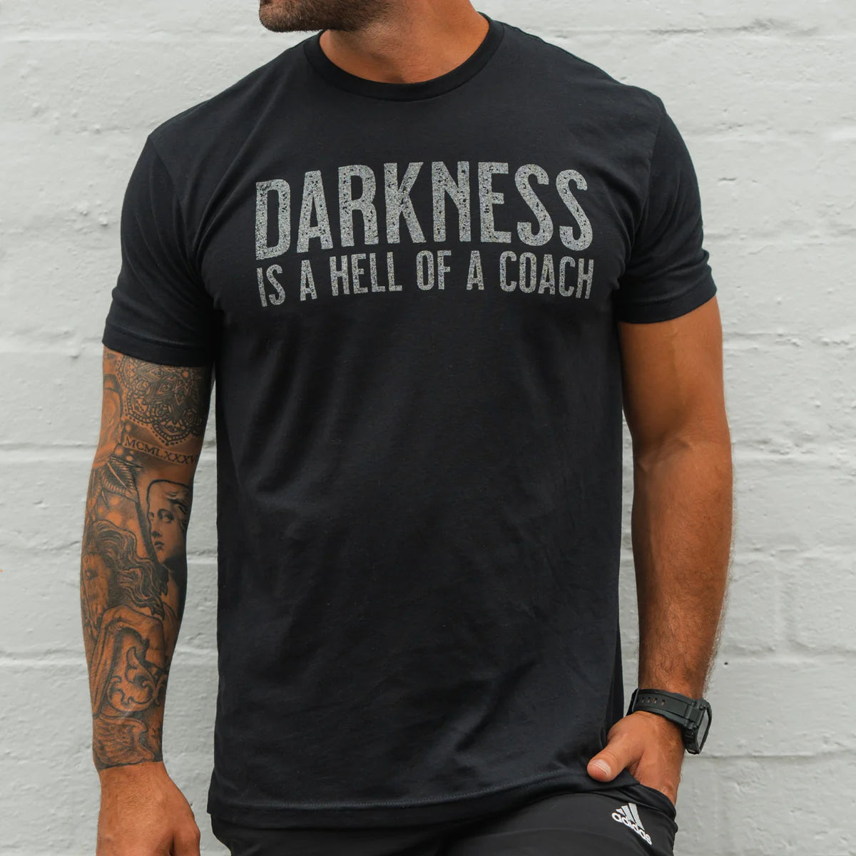 A man in the Darkness Is A Hell Of A Coach T-Shirt - Black by Tango Charlie Apparel, featuring bold white text, shows off his tattooed arm and watch against a light gray wall—embracing comfort and perfect stretch.