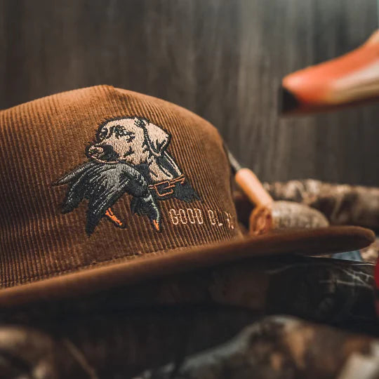The Mucker Bucket Good Ol Boy Corduroy Bucket is a brown corduroy hat with an embroidered dog carrying a duck, GOOD B-O-Y stitching, and a snapback for adjustable fit. It is shown against camouflage and a blurred wooden background.