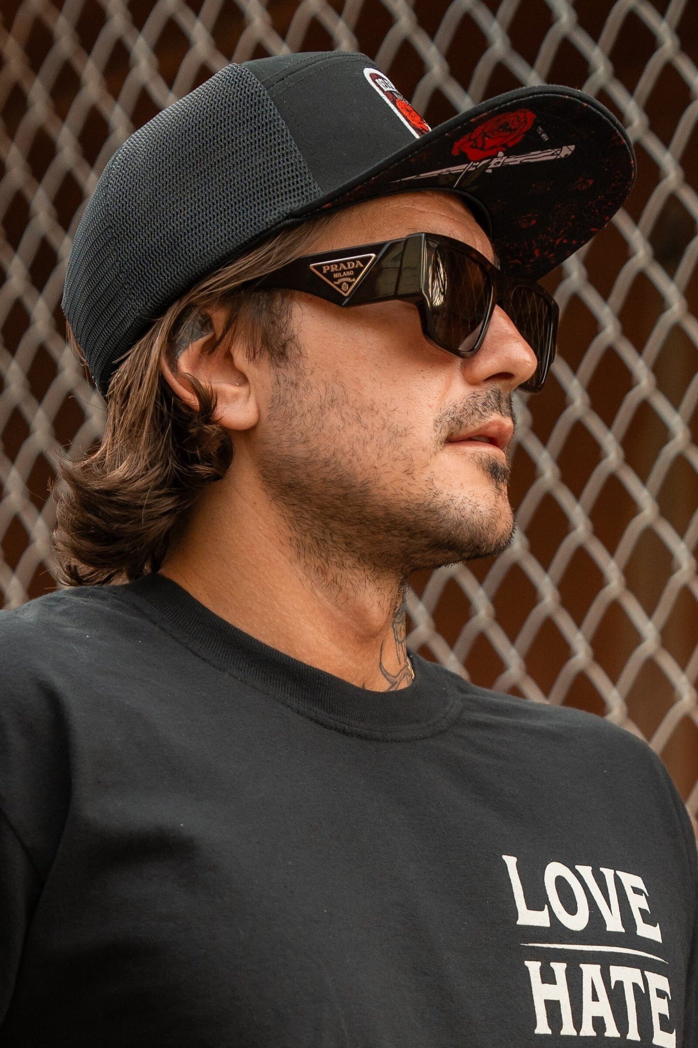 A person in front of a chain-link fence wears designer sunglasses and a black T-shirt with LOVE above HATE, paired with the Relentless Betrayal Love/Hate 7 Panel Trucker Snapback, highlighted by a patch with red and white details.